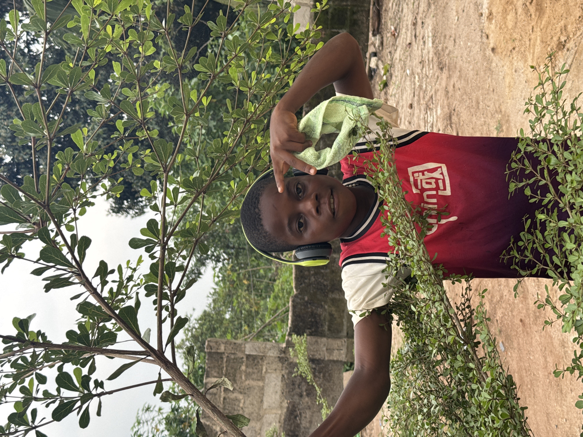 A young boy with dark skin and short dark hair smiles directly at the camera, wearing black and lime green over-ear headphones. He is dressed in a red and black short-sleeved t-shirt with white stripes and white sleeves. His left hand is raised, making a 'V' gesture, with a light green cloth draped over it.
The boy is positioned outdoors amidst lush green foliage, with branches and leaves partially obscuring him in the foreground and midground. Behind him, an unpaved, earthy ground is visible, along with a grey stone structure to his left and additional trees in the background, all under an overcast sky. The visible text on his t-shirt includes white Chinese characters, prominently featuring '国' (guó, meaning nation or country) and '一' (yī, meaning one), with another character partially obscured between them. The photograph captures a daytime scene, likely under cloudy conditions, in Eziudo, Nigeria.