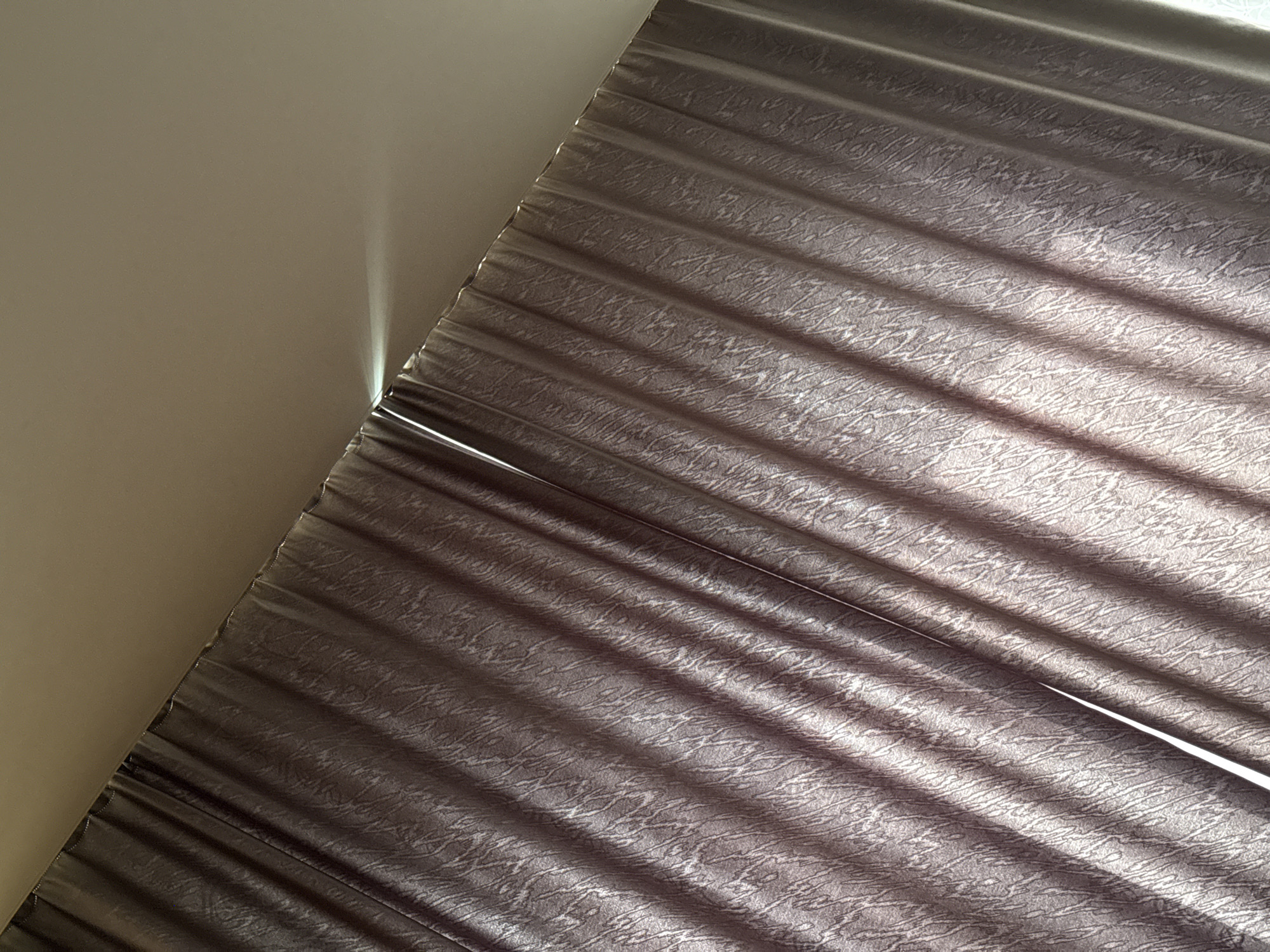The image presents an abstract indoor scene dominated by a textured window blind or curtain, illuminated by natural light. The composition is diagonal, with a plain, light beige or grey-brown wall/ceiling occupying the upper left portion and the pleated blind filling the lower right.
A prominent feature is the strong, directional sunlight entering the room. A vivid, narrow beam of bright white light cuts through a small gap between the top edge of the blind and the adjacent wall or ceiling, creating a striking highlight. This intense light suggests direct sunlight, indicating either early morning or late afternoon.
The blind itself is made of a medium-to-dark grey fabric, possibly with a subtle sheen, and features a distinct, intricate, abstract pattern resembling tangled lines or organic veins across its surface. The fabric is pleated or folded horizontally, creating depth. Parallel shadows are cast diagonally across the blind's surface, enhancing the texture and creating a striped effect that contrasts with the blind's own folds. These shadows vary in intensity, making certain areas of the blind appear darker than others, while illuminated sections show more of the intricate pattern.
There are no people, specific activities, or visible text depicted in the image. The scene focuses purely on the interplay of light, shadow, and texture within a domestic setting, highlighting the material qualities of the blind and the atmospheric effect of the sunlight.