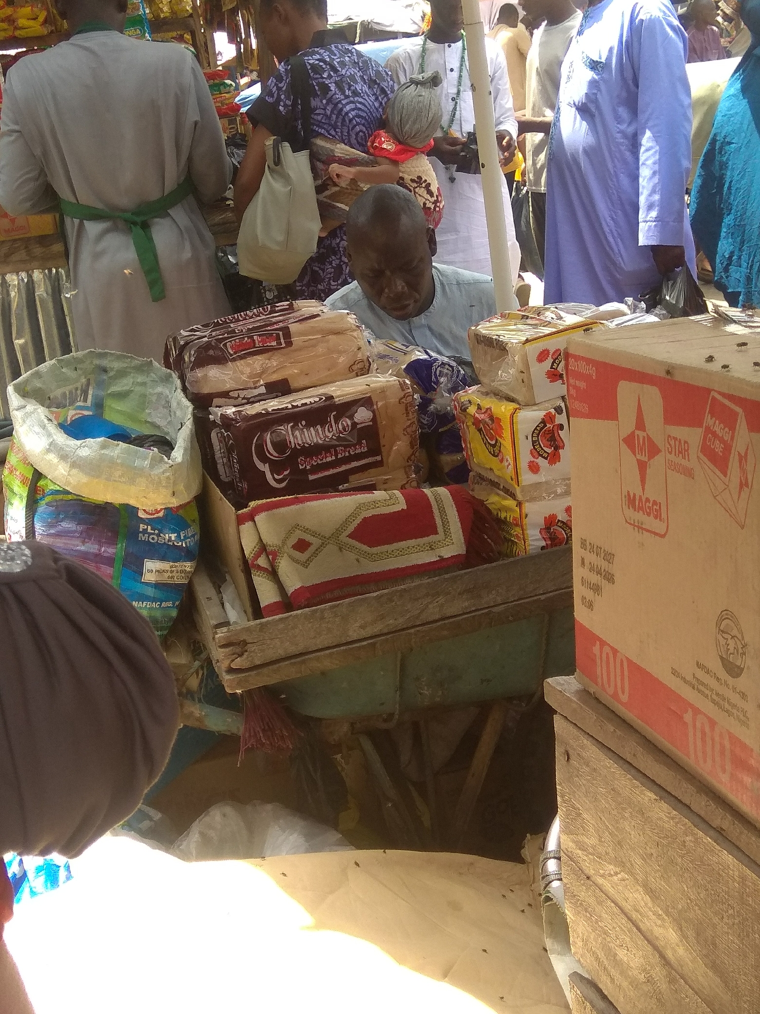The image depicts an outdoor market scene in Jalingo, Nigeria. In the foreground, a man wearing a blue shirt is partially visible, looking down. To his right, a large cardboard box labeled "MAGGI CUBE" is prominent, with the number "100" printed on it. Next to the box are stacks of bread loaves, some wrapped in plastic. To the left of the bread, a large open bag contains colorful items, possibly food or other goods. In the background, several people are visible, some appearing to be vendors and customers. They are dressed in various traditional and modern clothing. The market setting is suggested by the presence of goods for sale and people interacting. The overall impression is one of a bustling local market.