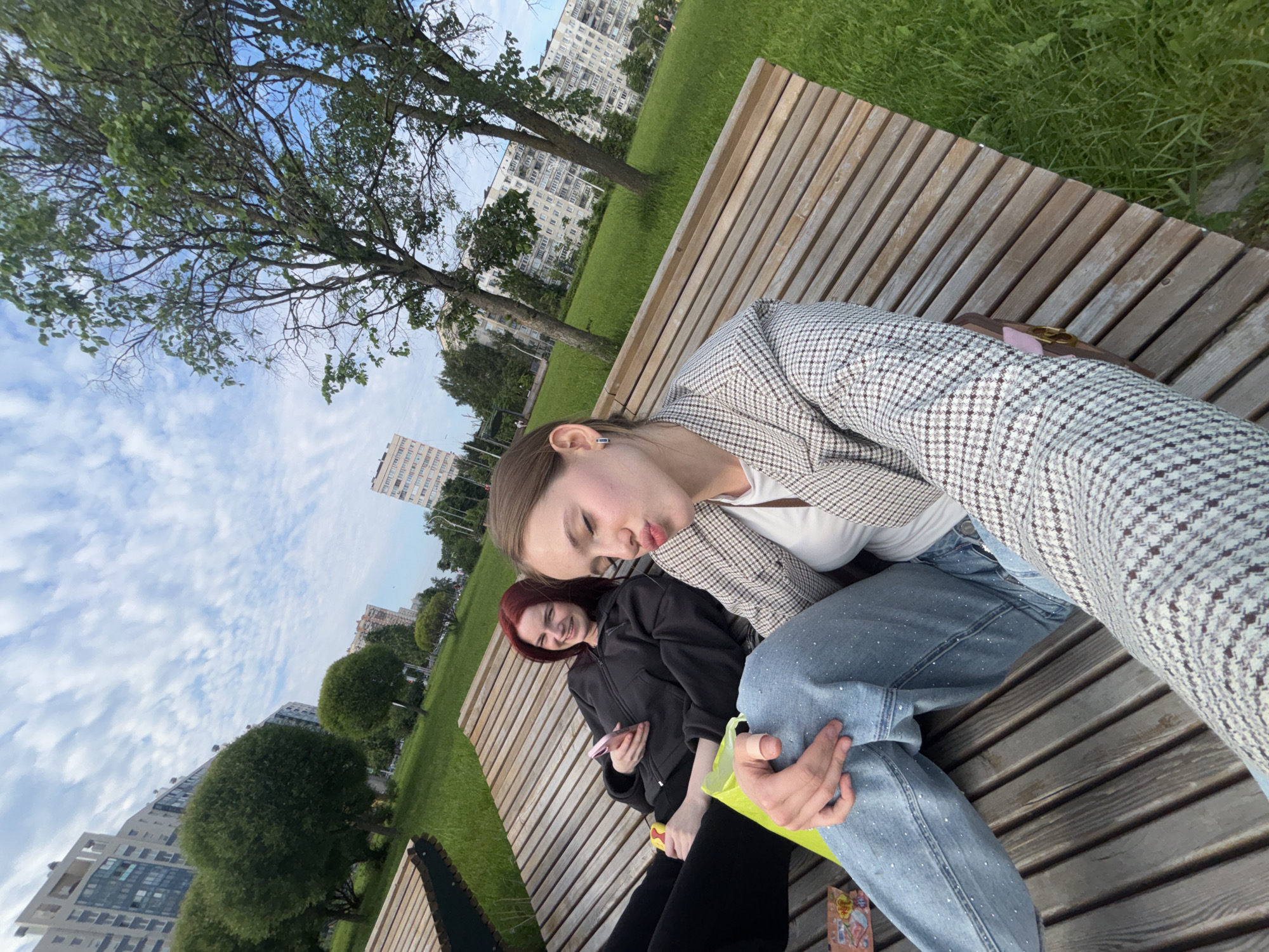 Two young women are sitting on a wooden park bench in a grassy urban park. The woman in the foreground is taking a selfie, puckering her lips. She is wearing a checkered blazer over a white top and studded blue jeans. Her right hand rests on her leg, and a small pink purse is visible beneath her. The woman in the background has bright red hair and is smiling at the camera. She is wearing a dark hoodie and black pants, and she is holding a pink smartphone in her left hand. A bright yellow shopping bag is on the bench between them. In the background, tall apartment buildings and trees line a green lawn under a cloudy sky. The scene is likely in the city of Sinyavino, Russia, given the architectural style of the buildings.