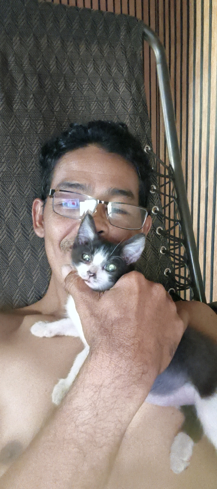 A selfie is taken from a person's perspective, showing their upper body and a small black and white kitten they are holding. The person is wearing glasses and has dark, curly hair. The kitten's face is close to the person's face, with its green eyes visible. The background consists of a dark, patterned fabric and a section of vertical wooden planks with dark lines, likely part of an interior or outdoor seating area. The person's chest and shoulder are visible, and their skin appears to have some texture and possibly hair. The scene appears to be indoors or in a sheltered outdoor space in Muaro Jambi, Indonesia, given the visual cues of the surroundings.