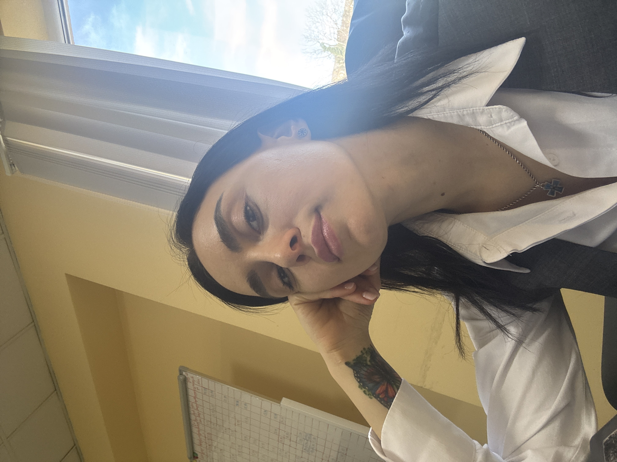 A woman with dark hair is resting her head on her hand, looking at the camera. She is wearing a white shirt and a gray vest. She is also wearing a necklace with a cross pendant. She has a tattoo of a butterfly on her wrist. The background is a yellow wall and a window with blinds. A whiteboard with writing is visible in the lower left corner of the image. The location is likely an office or workspace. It is not possible to ascertain that the location is specifically in the city of Tbilisi, Georgia, based on the visual evidence provided.