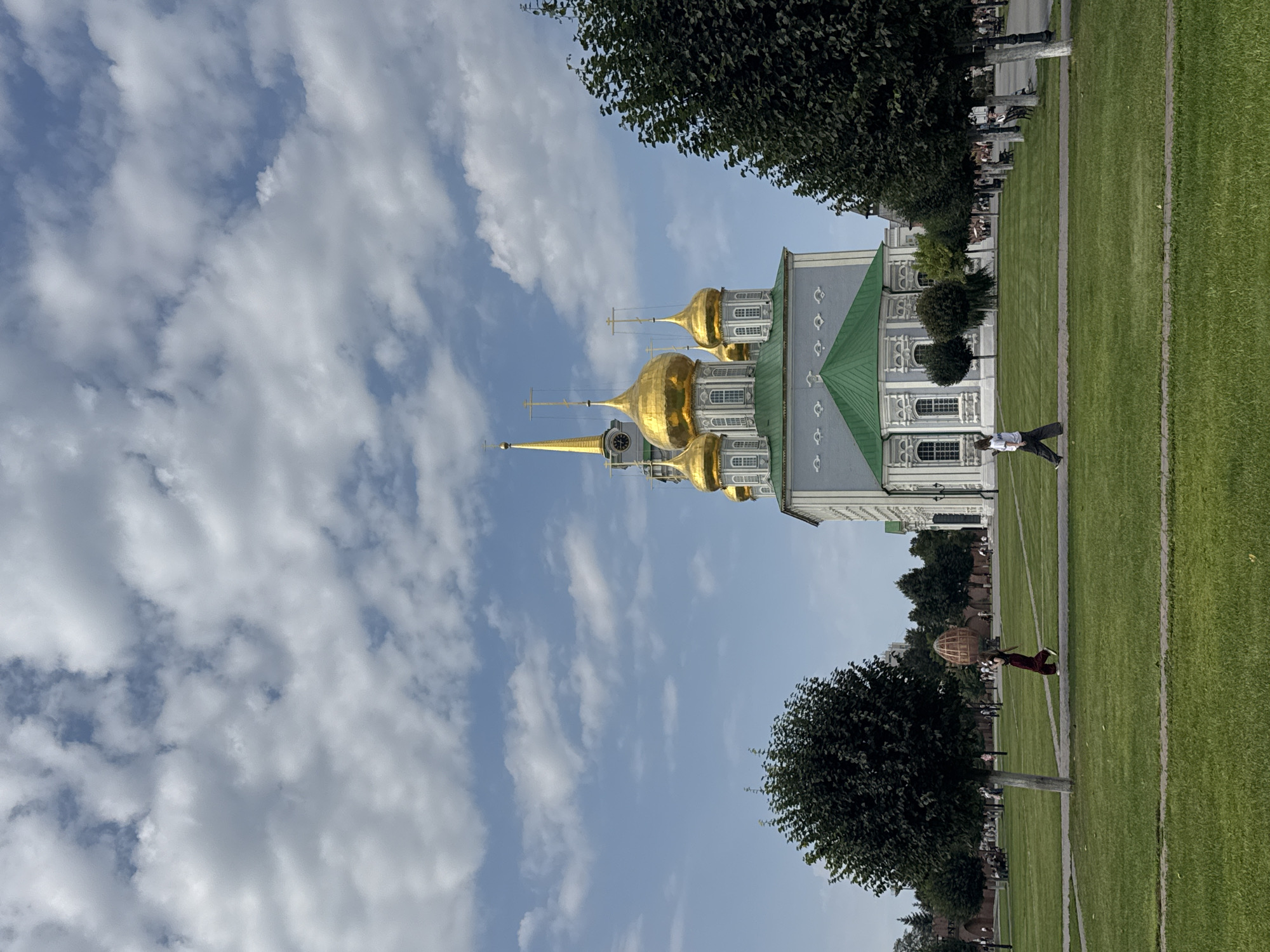 The image depicts the Assumption Cathedral in Tula, Russia, under a partly cloudy sky. The cathedral, a prominent landmark, features multiple golden domes and a tall, slender spire topped with a cross. Its facade is a pale blue-gray with white decorative elements and green accents. In the foreground, a neatly manicured green lawn extends toward a paved walkway. Two individuals are walking on the path; one in the mid-ground on the left is wearing a dark garment and a head covering, while the other on the right is dressed in a white shirt and dark pants. Trees line the walkway to the right of the cathedral and in the mid-ground on the left, providing shade and framing the architectural structure. In the background, more trees and the brick wall of the Tula Kremlin can be seen, with other people visible further in the distance, suggesting a public space.