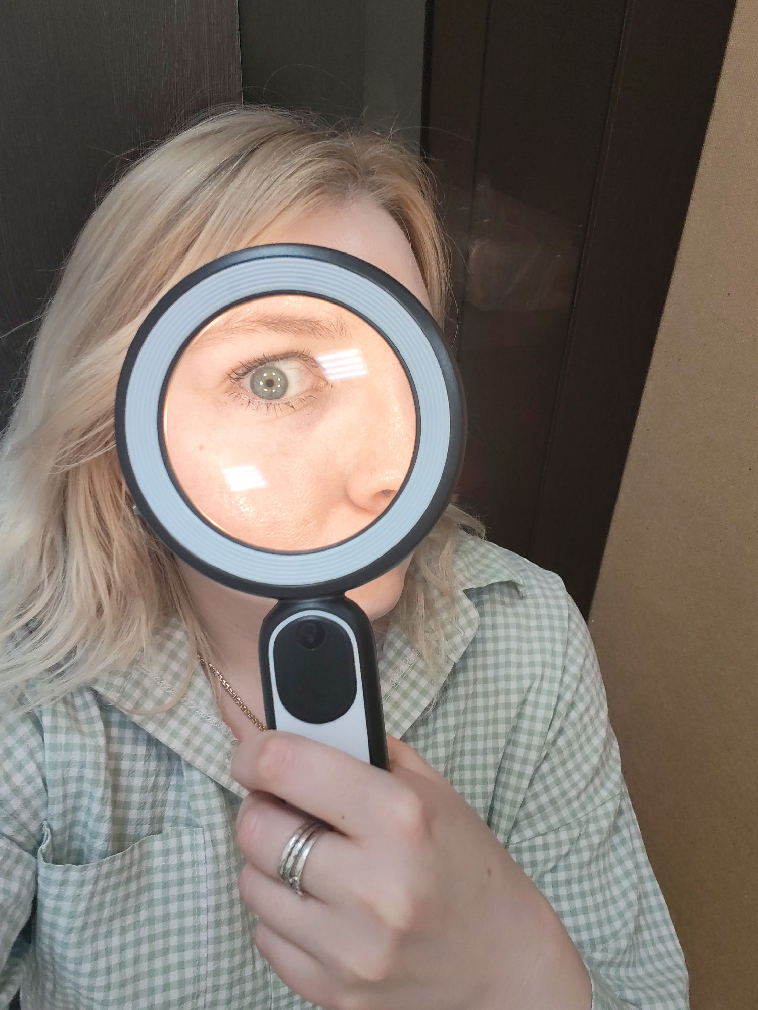 A person with fair skin and blonde hair is holding a magnifying glass up to their face, such that the glass frames their right eye and part of their nose and cheek. The person is wearing a light green and white checkered button-up shirt with a pocket visible on the left side. Three rings are visible on the ring finger of the hand holding the magnifying glass. A silver necklace is visible at the collar. The magnifying glass is black and white with a ring light illuminating the person's eye. The background consists of a dark brown wooden door or panel on the left and a light brown wall or panel on the right. There is no information available to indicate this image was taken in Izhevsk, Russia.