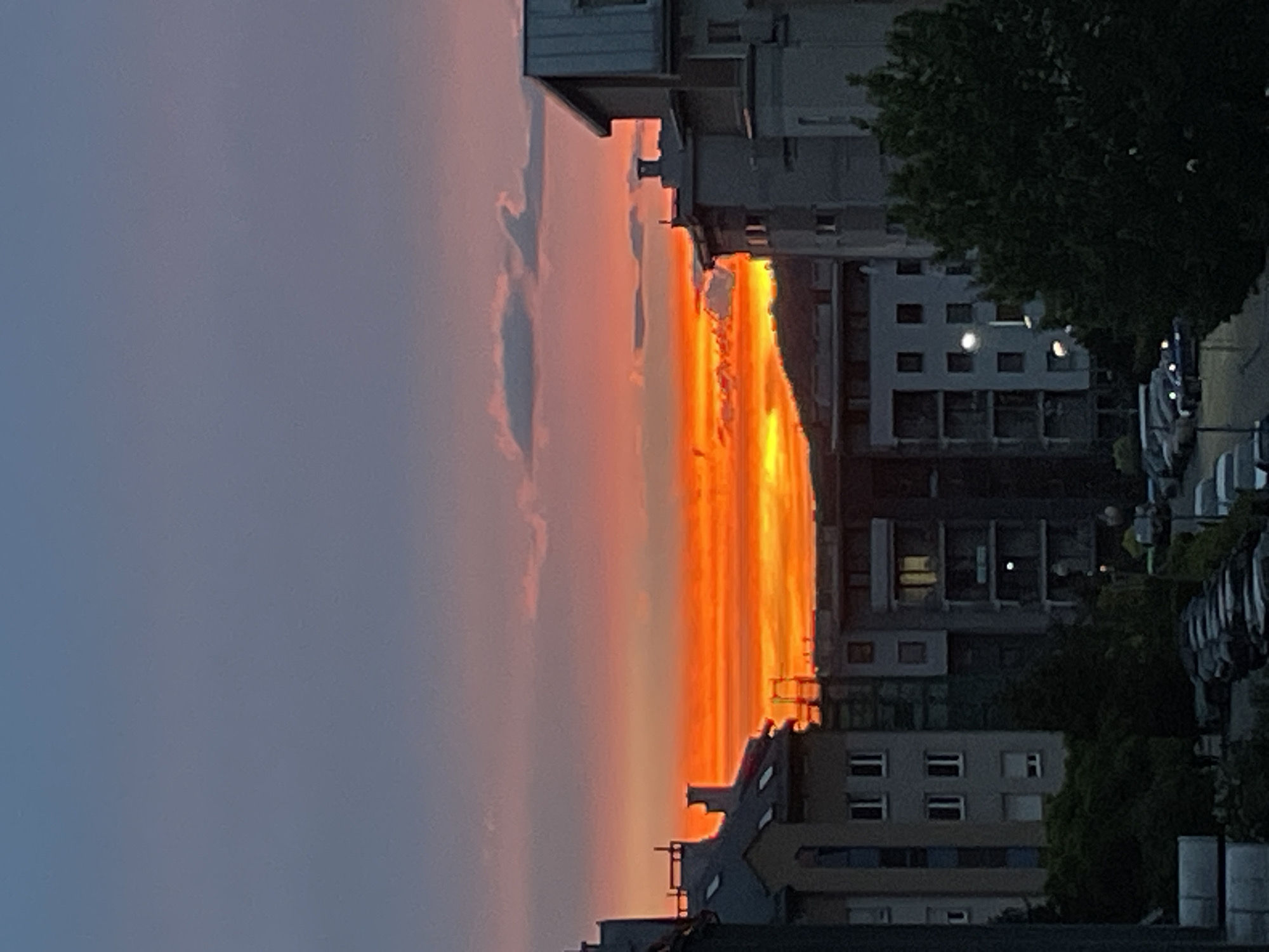 The image shows a vertical view of a cityscape during a vibrant sunset. An orange and yellow colored sky is framed by buildings on either side. The buildings on the right appear to be residential, with multiple windows visible. Trees are interspersed among the buildings. Cars are parked along the street on the right. The sky fades from orange and yellow near the horizon to a bluish-gray color higher up in the image. A few faint clouds are present in the sky.