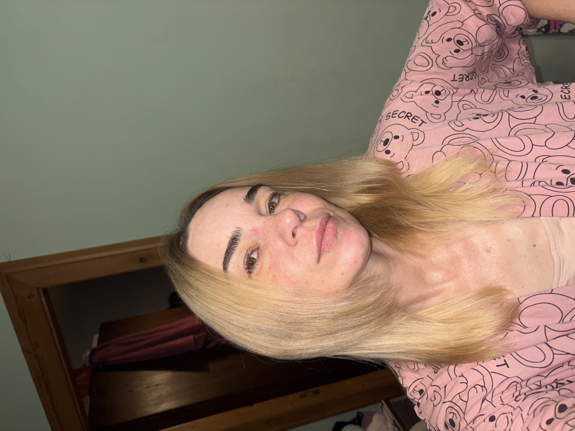 A medium shot shows a fair-skinned woman with blonde hair and light brown eyes. Her hair is shoulder-length with lighter blonde highlights. She has freckles on her face and some reddish blemishes. She is wearing a pink robe with a pattern of black teddy bears and the word "SECRET" repeated throughout. The background features a wooden doorway on the left, revealing a dark interior with some hanging clothes. The wall to the right is a light green color. The scene appears to be indoors, possibly in a bedroom.