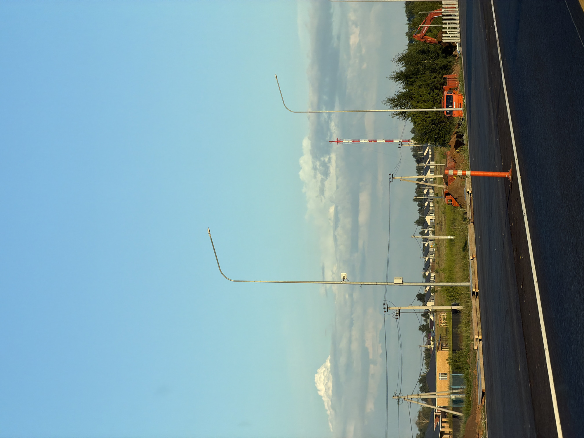 The image depicts a construction site on the outskirts of Kaipy, Russia, with a clear blue sky and scattered clouds. In the foreground, a freshly paved asphalt road with a white dividing line is visible. A bright orange traffic cone stands near the right edge of the road.
Further back, several modern houses with light-colored facades are lined up in a semi-rural setting, suggesting a developing residential area. Several utility poles with electrical wires crisscross the scene.
Dominating the middle ground are construction vehicles, including an orange truck and a red excavator, positioned near piles of dirt. A tall, red and white striped communication tower rises in the background. Green trees and shrubbery are interspersed throughout the landscape. The overall impression is one of ongoing infrastructure development in a newly developing area.
