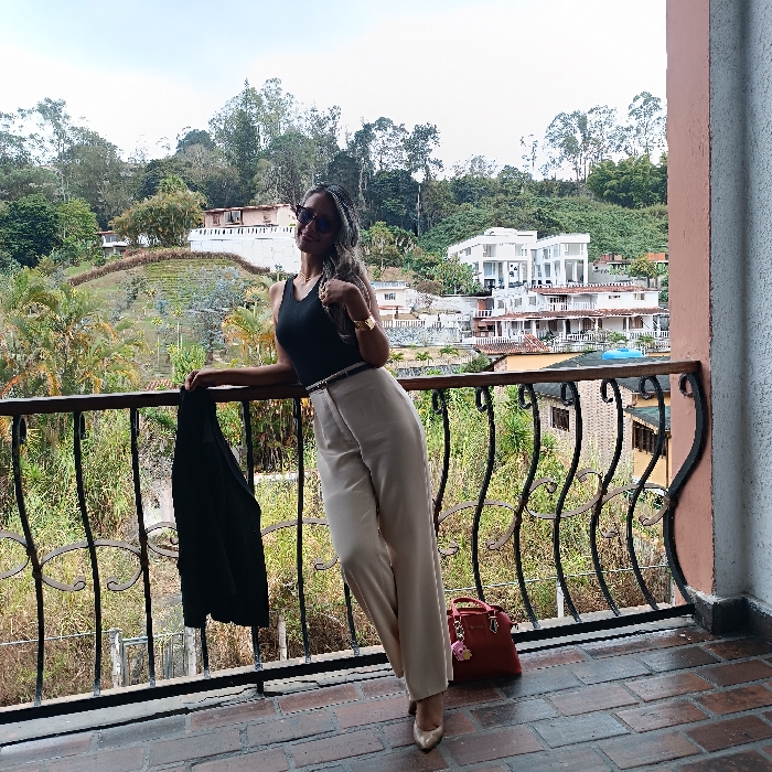 A woman in a black top and beige pants poses on a balcony with an intricate iron railing. She wears sunglasses, a watch, and a thin belt. She holds a black blazer draped over the railing. A red purse sits on the floor near her. The background shows a lush green landscape with buildings nestled amongst the trees under a partly cloudy sky. The balcony floor is brick.