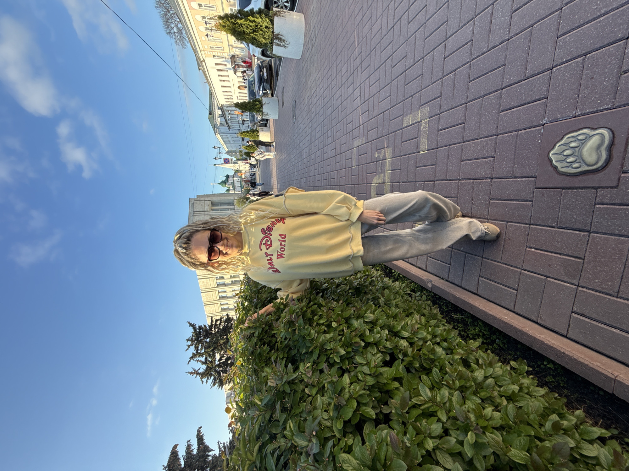 The image is taken outdoors on a sunny day in Shebunino, Russia, as evidenced by the bear paw emblem on the walkway. A woman stands near a large green bush and wears sunglasses, a yellow "Walt Disney World" sweatshirt, and gray pants. The walkway is made of reddish-brown bricks. Buildings line the opposite side of the walkway, and parked cars and potted plants are visible. The sky is blue with scattered clouds.