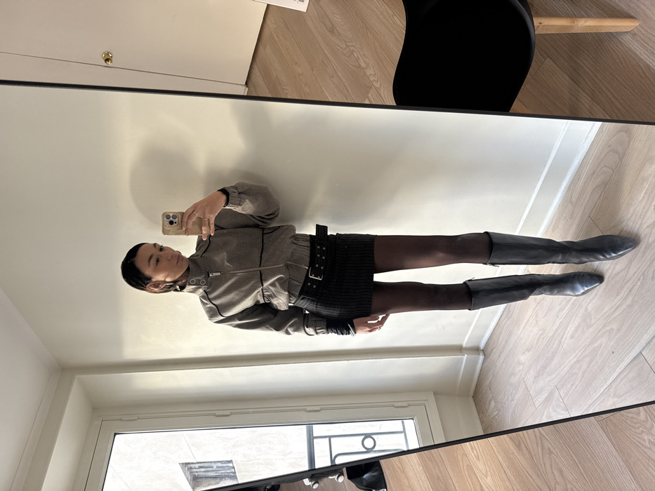 The image displays a full-body mirror selfie taken indoors, featuring a person, likely a woman, standing and capturing her reflection. The photo is vertically oriented, showing the individual from head to toe.
The person has dark hair, pulled back from her face, and fair skin. She holds a light-colored smartphone with a matching case in her right hand, positioned to take the selfie. Her gaze is directed towards the phone's camera, with a neutral expression.
She is dressed in a fashionable ensemble. Her top is a light grey or beige jacket, possibly with a subtle textured pattern like a micro-houndstooth or fine stripes. It features a high collar, a central zipper with black trim details, and elasticated cuffs and hem, giving it a modern, slightly casual yet structured look. Below the jacket, she wears a short, dark-colored skirt, likely black, which has a distinct ribbed or pleated texture. A prominent black belt with a large, rectangular silver-toned buckle and several metal eyelets is cinched around her waist, adding an edgy detail to the outfit. Her legs are covered in dark, opaque tights or pantyhose, which extend into a pair of dark, possibly black or charcoal grey, knee-high boots with a subtly pointed toe. A ring is visible on one of her fingers on the hand holding the phone.
The background reflected in the mirror reveals a clean, well-lit interior. The wall directly behind the person in the reflection is a plain white or off-white. On the left side of the reflection (which is actually behind the person's right side), a white door with a round brass doorknob is partially visible. On the right side of the reflection (behind her left side), a dark piece of furniture, possibly a chair, with a light wooden leg is partially seen. The floor in the reflection is made of light-colored wooden planks. Below the person's feet, the mirror reflects a window, offering a glimpse of an outdoor scene, possibly a building or courtyard, suggesting the room is on an upper floor.
The lighting appears to be bright and even, likely a combination of natural light from the window and ambient indoor light, creating soft shadows on the wall. The overall impression is a personal moment captured, focusing on the individual's outfit and style. No discernible text is visible within the image.