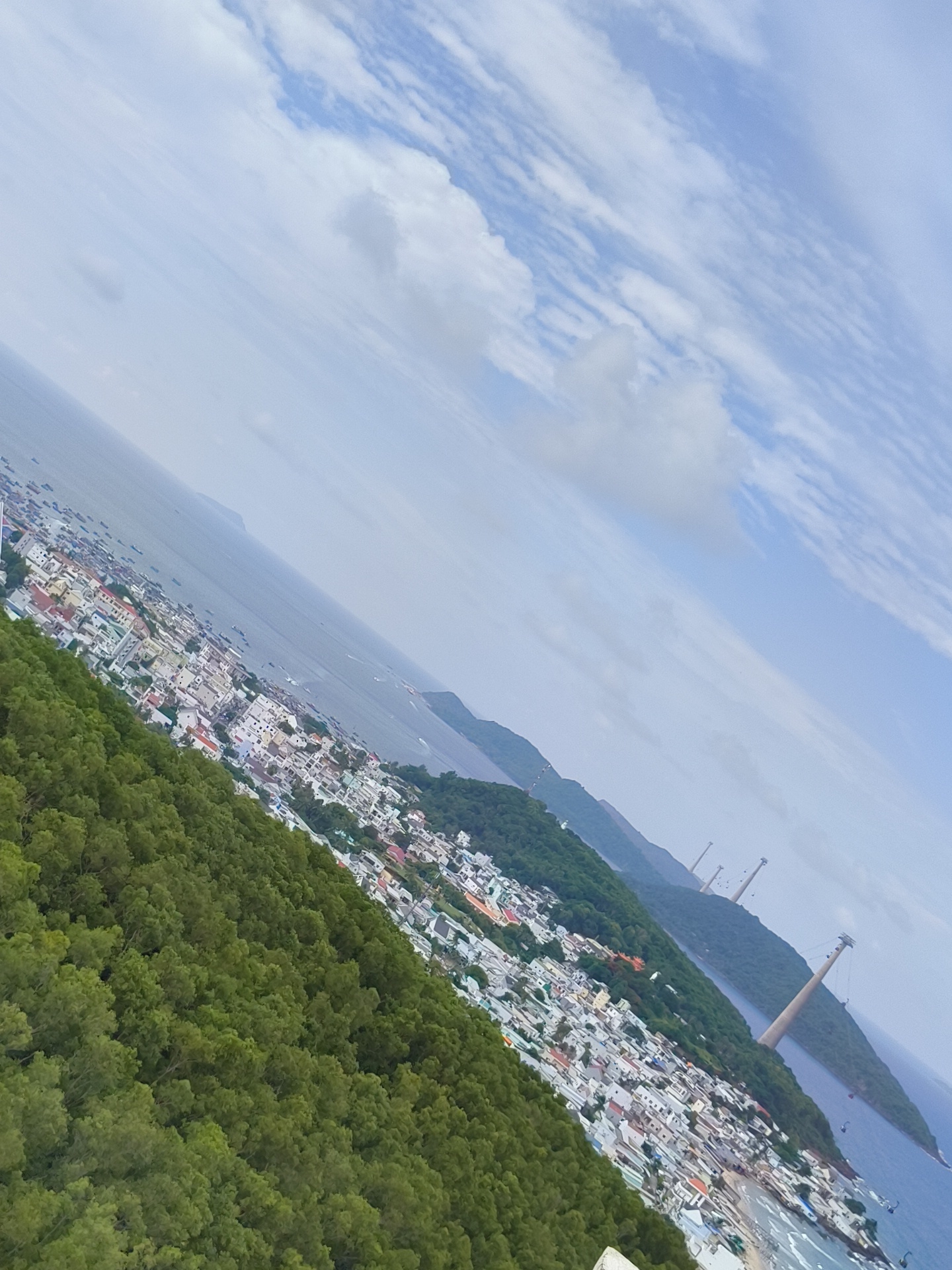 The image is a high-angle shot of the city of Phú Quốc, Vietnam, as viewed from a wooded hillside. The city is nestled along the coast, with a dense collection of white buildings with red-tiled roofs. The sea is visible in the background, dotted with numerous small boats. In the distance, three towering cable car pylons extend from the sea onto a verdant, mountainous island. The sky above is partly cloudy, with streaks of white clouds against a pale blue backdrop. The overall scene conveys a sense of a coastal urban environment bordered by lush natural landscapes.