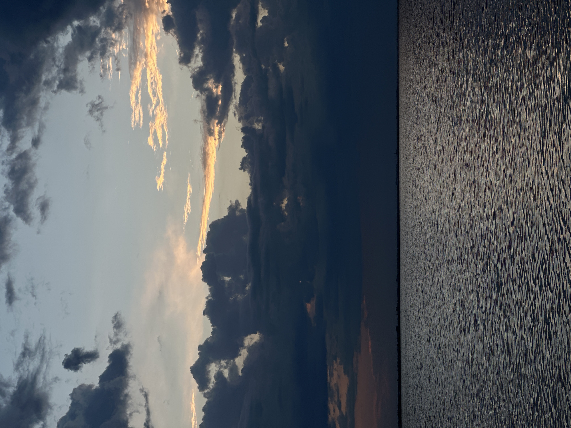 The image captures a serene, expansive view of a body of water under a dramatic sky. The water, appearing vast and calm, reflects the subdued light of either dusk or dawn. The surface is textured with gentle ripples, creating a shimmering effect. In the distance, a thin, dark horizon line suggests land, possibly the distant shore of a lake or sea, with faint silhouettes of what might be structures or trees barely discernible against the dim sky.
The sky is a canvas of transitioning light and shadow. Large, dark, and brooding clouds dominate the upper portion of the image, their forms suggesting depth and an impending change in weather. Interspersed between these darker masses are streaks and patches of soft, warm light, primarily golden and pale orange hues, indicating where the sun is either rising or setting, breaking through the cloud cover. The overall atmosphere is one of twilight tranquility, with a hint of dramatic intensity due to the contrasting light and cloud formations.
This scene is evocative of the natural beauty found in and around Kokshetau, Kazakhstan, a city known for its proximity to numerous lakes, including the prominent Lake Borovoye and the larger Lake Shchuchye, which are part of the Burabay National Park. The vast expanse of water and the characteristic steppe sky with its dramatic cloudscapes are characteristic of the landscapes in this region. The image does not contain any people or specific identifiable objects that would pinpoint a precise location within the city, but it strongly represents the type of natural scenery that defines the Kokshetau area, especially its lakeside vistas during twilight hours.