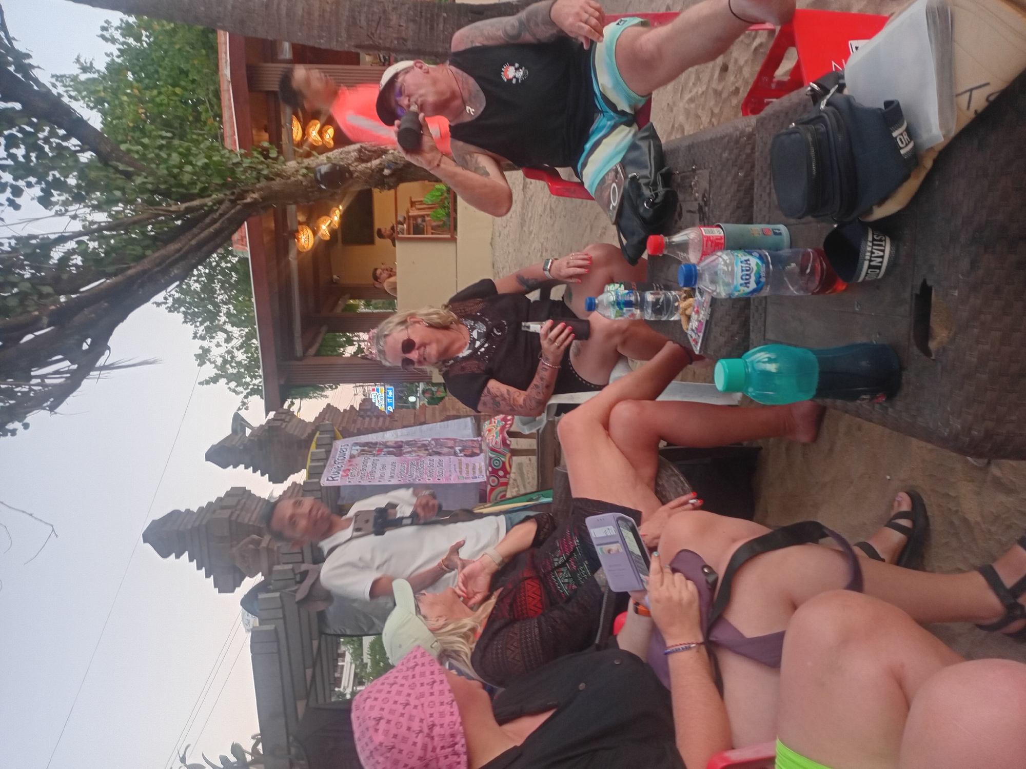 In Kuta, Indonesia, a group of individuals is gathered on a beach. The scene unfolds outdoors, with sand visible on the ground and a palm tree and lush foliage contributing to the tropical ambiance. In the background, a building with an open-air structure and ambient lighting suggests a bar or restaurant setting.
Several people are seated around a low-lying table. A woman in a pink knitted hat is on the left, looking down at her phone or tablet. Next to her, a woman wearing a light-colored cap and a dark patterned top is holding a bottle and appears to be engaged in conversation. On the right side of the table, a man wearing a black tank top and patterned shorts is drinking from a bottle. Another person, wearing a bright orange shirt, is partially visible in the background. A man in a white polo shirt, holding a camera or phone on a tripod, stands behind the seated individuals.
On the table, various objects are arranged, including bottles of water and what appears to be a soft drink, a small black bag, and some papers. The presence of beachgoers, a table setting, and recreational drinks strongly suggests a casual social gathering at a beachside establishment.