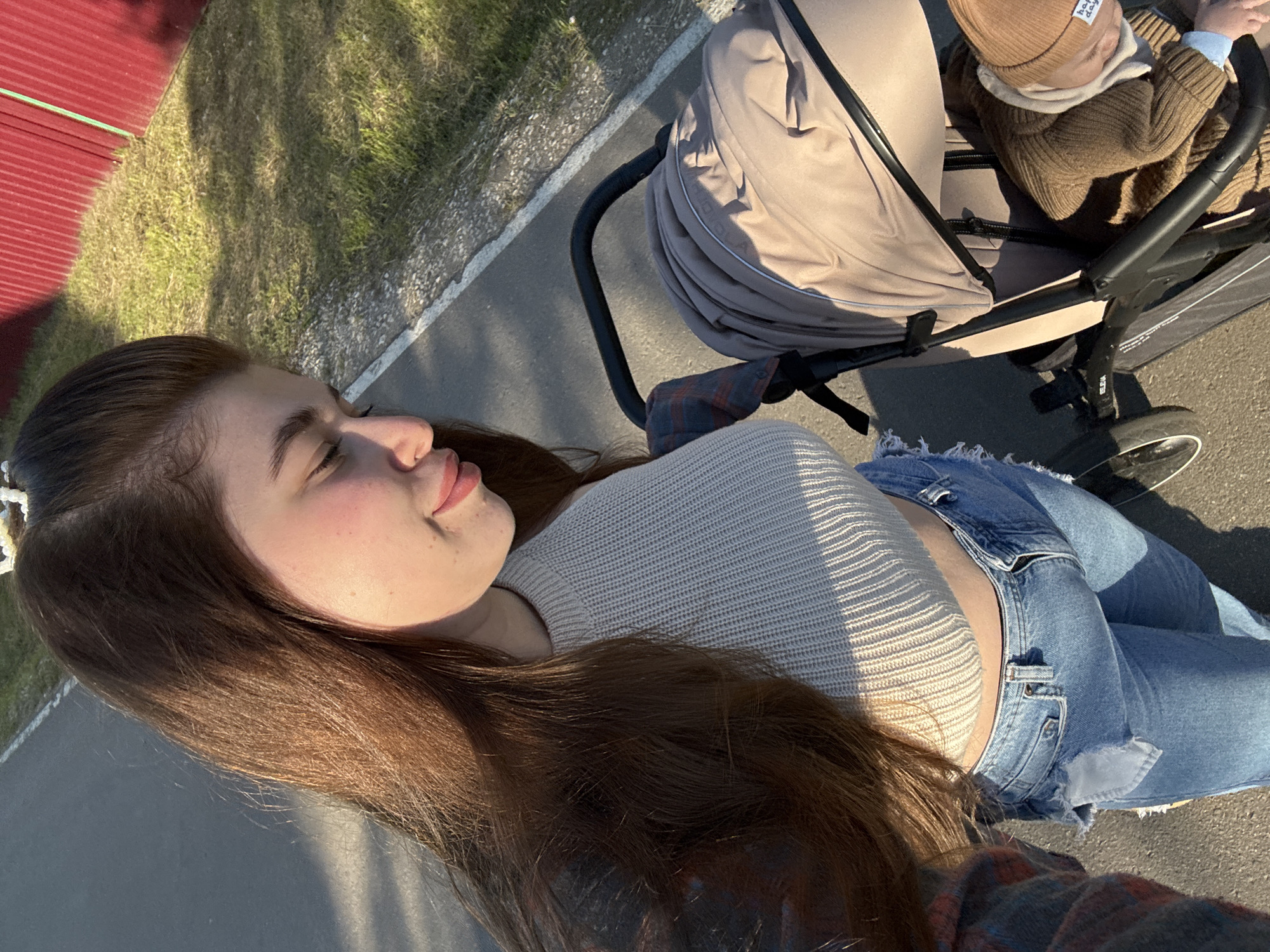 The image captures an outdoor scene featuring a young woman and a child in a stroller. The woman, viewed from a side profile, has long brown hair and wears a light-colored, ribbed long-sleeved crop top and blue denim jeans. Her eyes are closed or partially closed, and she has a gentle, serene expression.
Beside her is a beige baby stroller, holding a child who is partially visible from the back. The child is dressed in a brown knitted beanie with "happy day" written on it, a light-colored hooded layer, and a brown knitted cardigan. The stroller's frame, near the wheel, displays the text "ELENA". A plaid shirt is draped over the stroller's handle.
The setting consists of a paved path or sidewalk, bordered by green grass. In the background, a red metal fence or wall is visible. Strong sunlight casts clear shadows, indicating a bright, sunny day, likely during the late afternoon or early morning. The overall scene suggests a leisurely outdoor walk or outing. The location context provided is Мещёры, Russia.