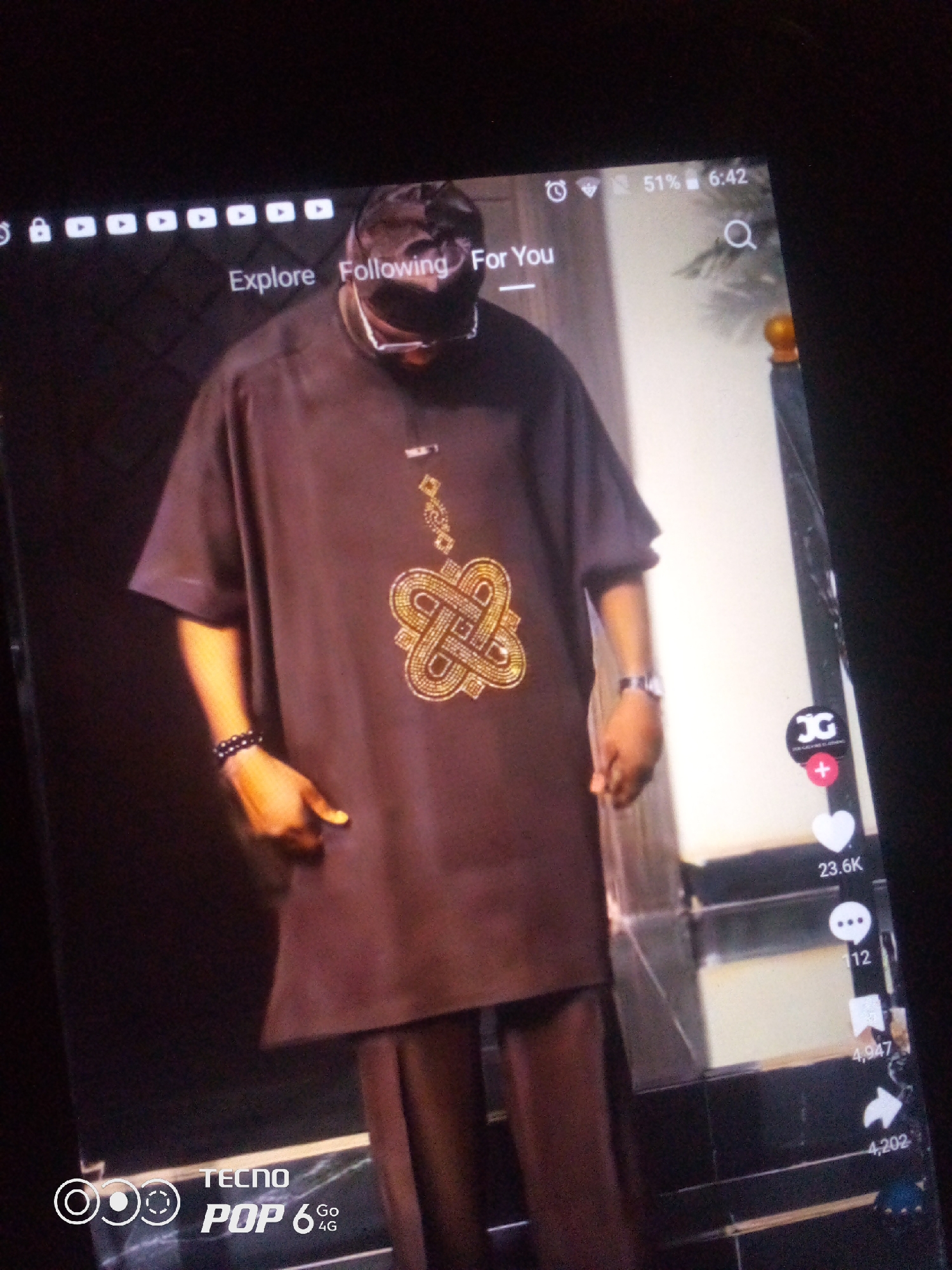 The image is a screenshot of a mobile device displaying a video on a social media platform. The video features a man wearing a dark brown, oversized tunic with intricate gold embroidery on the chest and a matching pair of trousers. He also wears a dark cap and eyeglasses. The man has bracelets on his left wrist. The interface of the mobile device is visible, showing icons for "Explore," "Following," and "For You" at the top. Below the video, social media interaction icons and engagement counts (e.g., 23.6K likes, 112 comments, 4,947 shares, 4,202 views) are displayed on the right side. The bottom left corner of the screen shows the "TECNO POP 6 GO 4G" branding, indicating the device model. The background of the video appears to be an indoor setting with reflective surfaces, possibly a studio or a well-lit interior. The specific location in the city of Howayi, Nigeria, cannot be determined from the image itself.