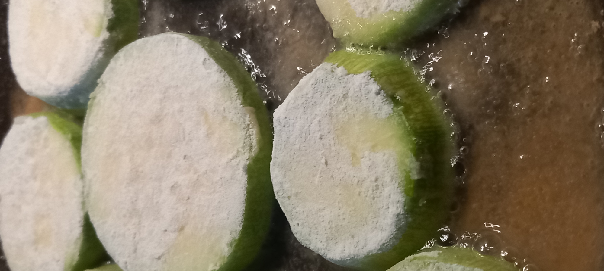 The image displays multiple thick, circular slices of a green vegetable, consistent with zucchini, being fried in hot oil within a pan or skillet. Each vegetable slice is coated uniformly with a white, powdery substance, indicative of flour or breading. The slices are partially submerged in a golden-brown liquid identified as cooking oil, which exhibits active bubbling around the edges of the vegetables and across the visible surface, indicating a high temperature. The underlying cooking surface is dark and indistinct. The environment is a kitchen setting in the city of Kryvyi Rih, Ukraine. No individuals are visible.