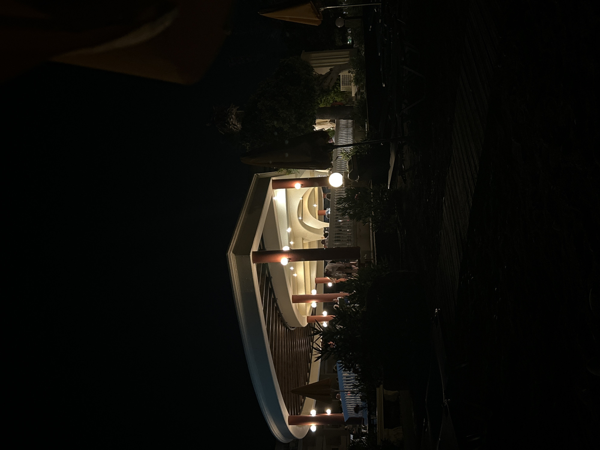 The image captures an outdoor seating area of a establishment at night, possibly a restaurant or bar, in Rhodes, Greece. A curved, covered structure with pillars illuminated by warm lights provides shelter and seating. Several people are visible in the background, some standing and others seated, suggesting a social gathering. Umbrellas, lounge chairs, and plants are scattered throughout the scene. The foreground is predominantly dark, with the light from the establishment casting some illumination on the paved ground. The overall ambiance is one of a relaxed evening setting.