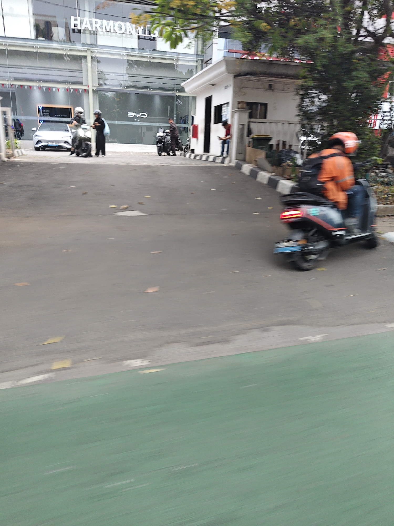 This is a street-level, medium shot, outdoor image taken in West Jakarta, Indonesia. The image captures a scene in front of a modern building with large glass windows and the prominent text "HARMONYLS" displayed above the entrance. In front of the building, there is a road with a green bike lane visible on the bottom. Several motorcycles and a white car are parked or in motion.
On the left side of the image, a white car is parked. Next to it, two individuals on a motorcycle, wearing helmets, are about to disembark or have just disembarked. Another motorcycle is parked further to the right. In the middle ground, a person in an orange jacket and a helmet is riding a dark-colored electric scooter, which is in motion and appears to be blurring due to the camera's movement or the scooter's speed. The scooter has its brake lights illuminated.
The environment appears to be an urban setting, possibly a dealership or a commercial complex, with trees and greenery visible behind the building. The overall impression is one of a bustling street scene with a mix of vehicles and pedestrians.