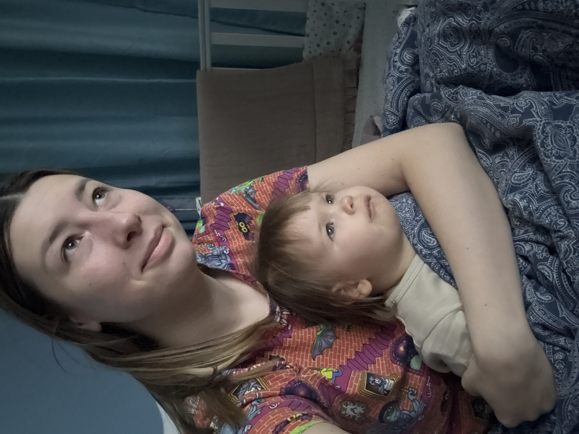 This indoor portrait features a young adult woman and a small child, likely a toddler, positioned closely together. The image is tilted, giving a dynamic perspective. The woman is lying down, supporting the child who rests on her arm or chest. Both subjects are looking upwards and to the right side of the frame, suggesting they are observing something above or beyond the camera's view.
The woman has light brown hair and is wearing a vibrantly patterned top featuring cartoonish monster characters, bats, brick walls, and framed pictures in shades of orange, purple, green, and blue. Her expression is gentle and somewhat pensive. The child, with light brown hair and blue eyes, appears calm and observant. They are wearing a simple, light-colored (possibly beige or cream) top.
The setting appears to be a bedroom. A dark blue duvet or blanket with an intricate paisley pattern covers part of the bed. In the background, muted teal curtains are visible, along with what looks like a light-colored headboard or part of a bed frame/crib, and a brown upholstered object which could be a pillow or part of the furniture. The lighting is soft and diffuse, typical of an indoor setting, and does not strongly indicate a specific time of day.
There are no visible texts or specific location cues within the image to confirm the provided context of Moscow, Russia. The overall mood of the image conveys a sense of quiet companionship and shared observation between the woman and child.