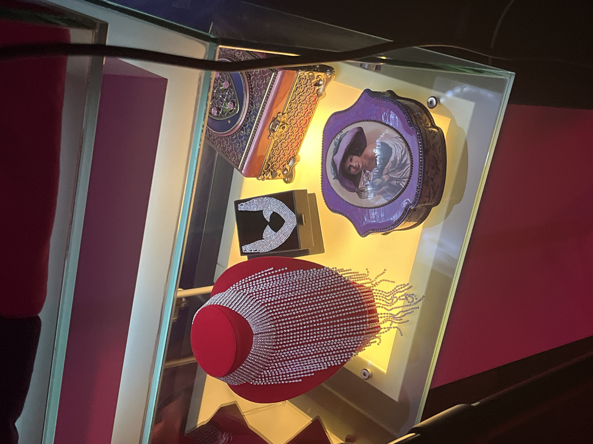The image displays a glass-encased display case with several items of jewelry and decorative boxes. In the center of the display, a red velvet bust showcases a long, cascading diamond-like fringe necklace. To the right of the bust, a black box holds a heart-shaped diamond necklace. Further to the right, a decorative wooden box with ornate gold trim and floral patterns sits atop a lower shelf. In front of the wooden box, a purple decorative box features a framed image of a woman wearing a wide-brimmed hat and a white garment. The display case is illuminated from below, casting a warm glow on the items. The background appears to be a dark, possibly red, fabric surface. Based on the visual evidence, it is not possible to determine the location within the city of Kazan, Russia. The scene depicts a static display of luxury items.