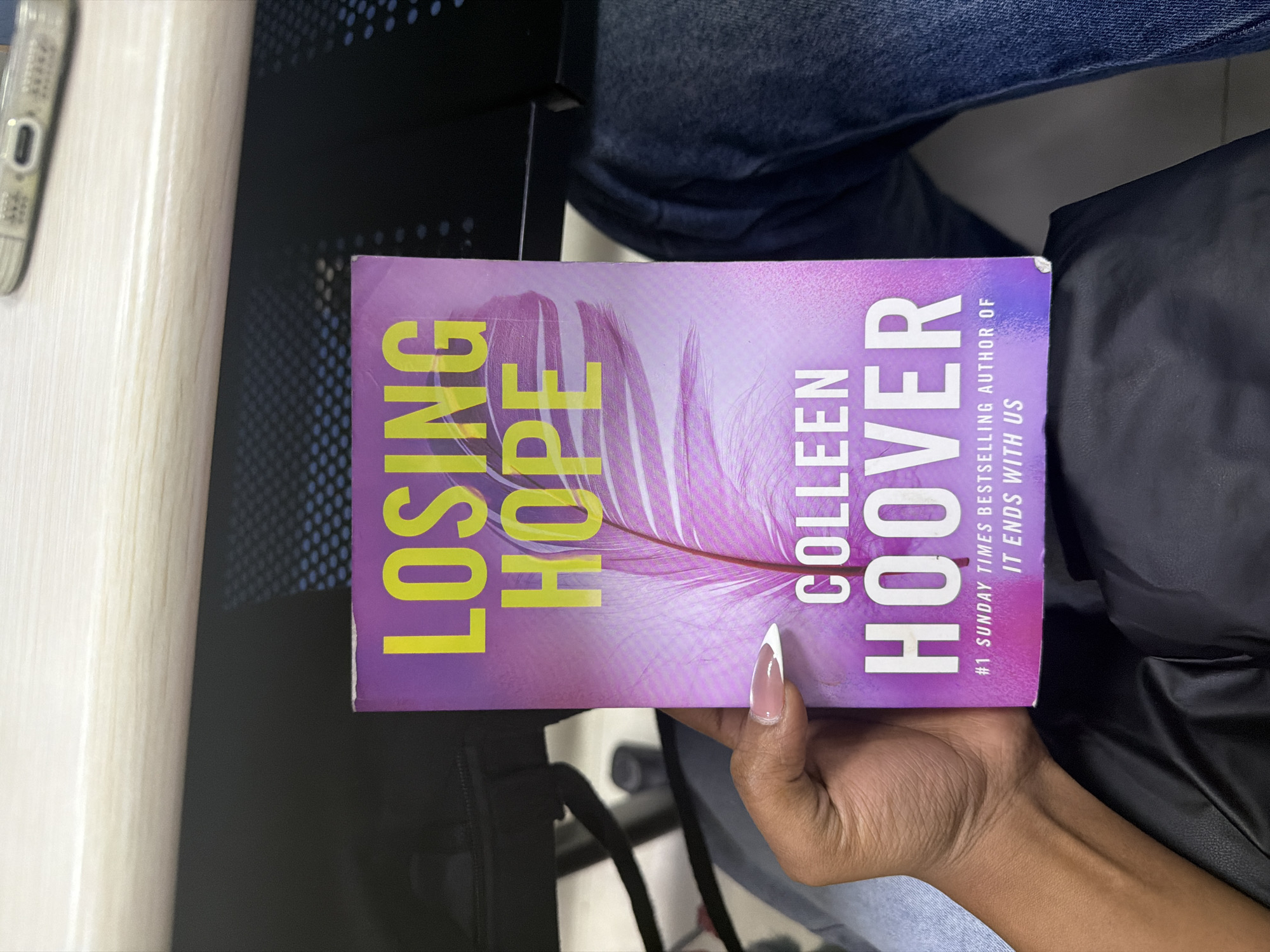 The image displays a book titled "Losing Hope" by Colleen Hoover held by a dark-skinned hand with a manicured nail. The book has a purple cover with yellow text and a feather graphic. The background includes a black chair with a mesh back, a light-colored surface with an object that could be a phone, a person wearing blue jeans, and a black jacket. The location or city is not discernible from the image.