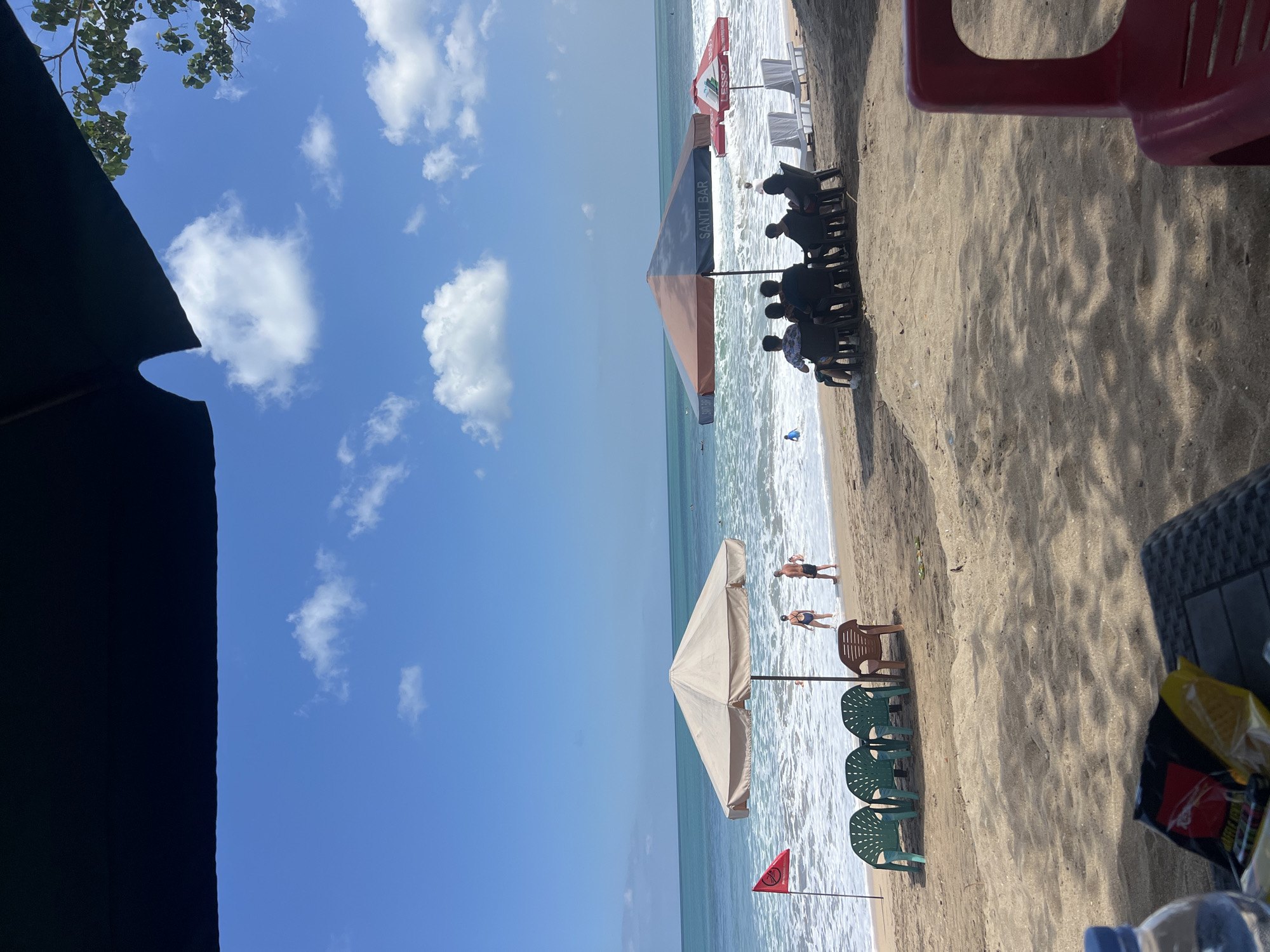This is a medium shot of a beach scene in Kuta, Indonesia. The foreground shows a sandy area with a red plastic chair on the right and a table with a water bottle and snacks on the left. The middle ground features a beach with several people, umbrellas, and chairs. Two people are standing in the shallow water near a red flag planted in the sand. Another group of people are seated under a large umbrella with "SANTI BAR" written on it. Beyond them, the ocean stretches out to the horizon, with small waves breaking on the shore. The sky is blue with scattered white clouds.