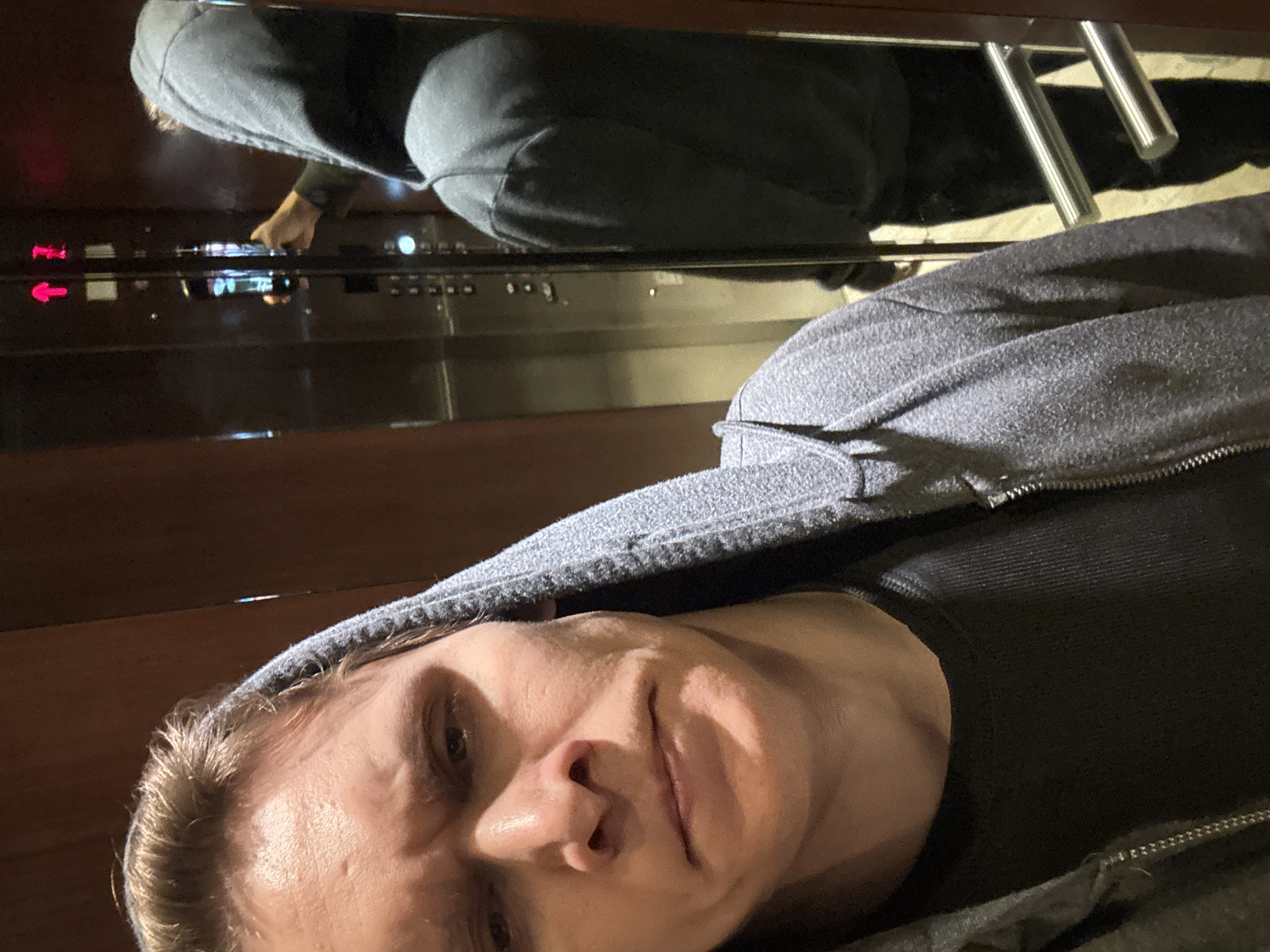 In an elevator, a man wearing a gray hooded jacket and a dark t-shirt takes a selfie. The elevator doors are made of mirrored stainless steel, reflecting the man and the elevator interior. The man's face is close to the camera, with a serious expression. He is holding a smartphone, which is partially visible in the reflection as he takes the photo. The elevator walls are a dark wood paneling. The image is grainy and appears to be taken in low light.