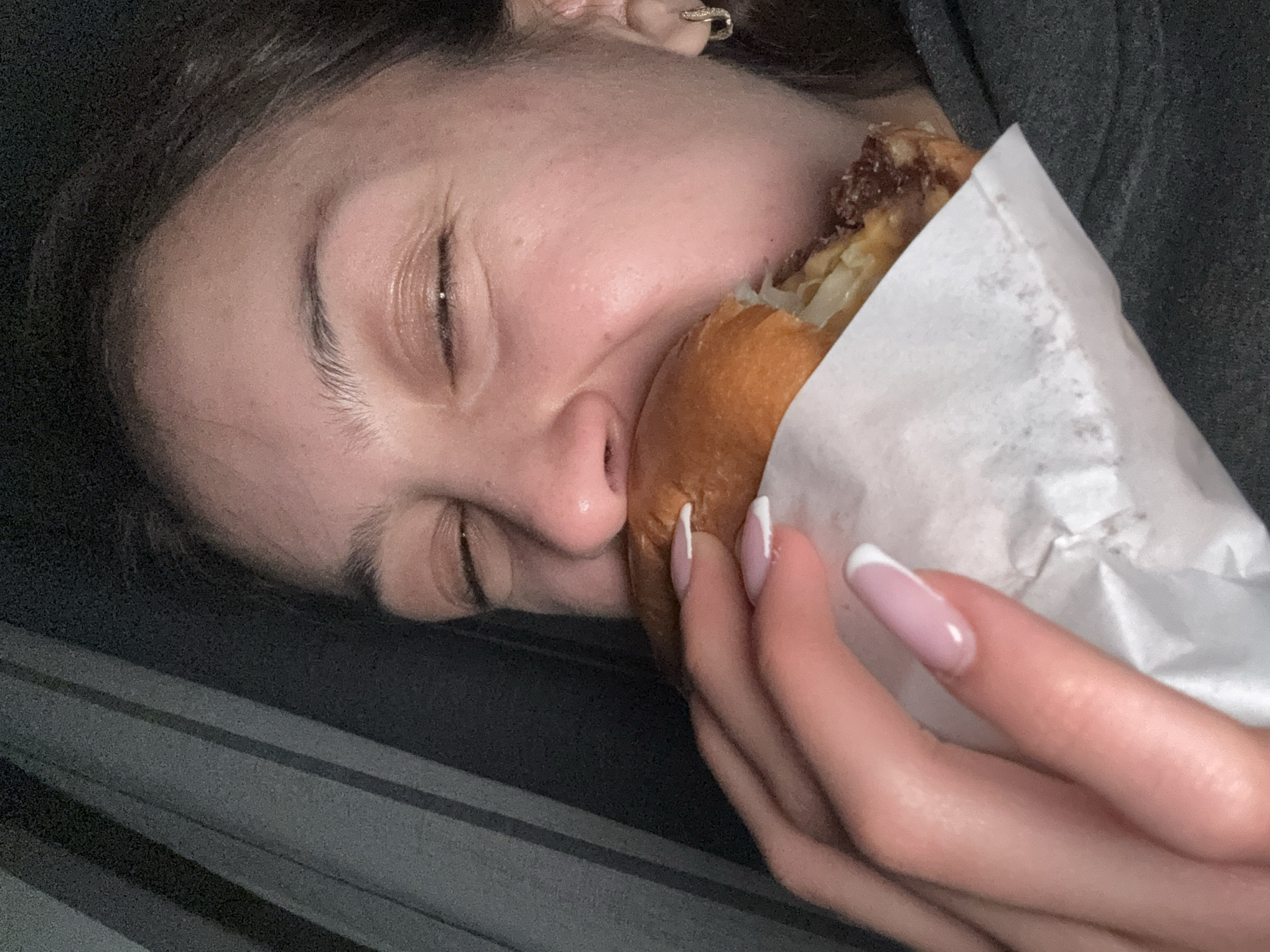 A close-up image features a person with fair skin and brown hair, lying on a surface, possibly a bed or couch. Their eyes are closed, and they are in the process of eating a large sandwich, held in a white paper wrapper. The sandwich appears to contain meat and other fillings. The person has manicured nails with a French tip design. They are wearing a small, possibly gold, earring in their visible ear. The overall environment is dark and could be interpreted as an indoor setting. There is no explicit evidence to suggest the location is Milan, Italy.