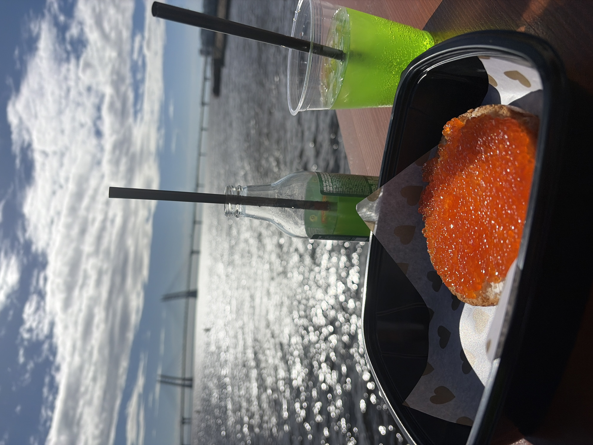 In Saint Petersburg, Russia, on a table by the water, a black container holds a piece of bread topped with red caviar. Beside it, a clear plastic cup and a glass bottle contain green beverages with straws. In the background, a bridge spans the shimmering water under a cloudy sky.