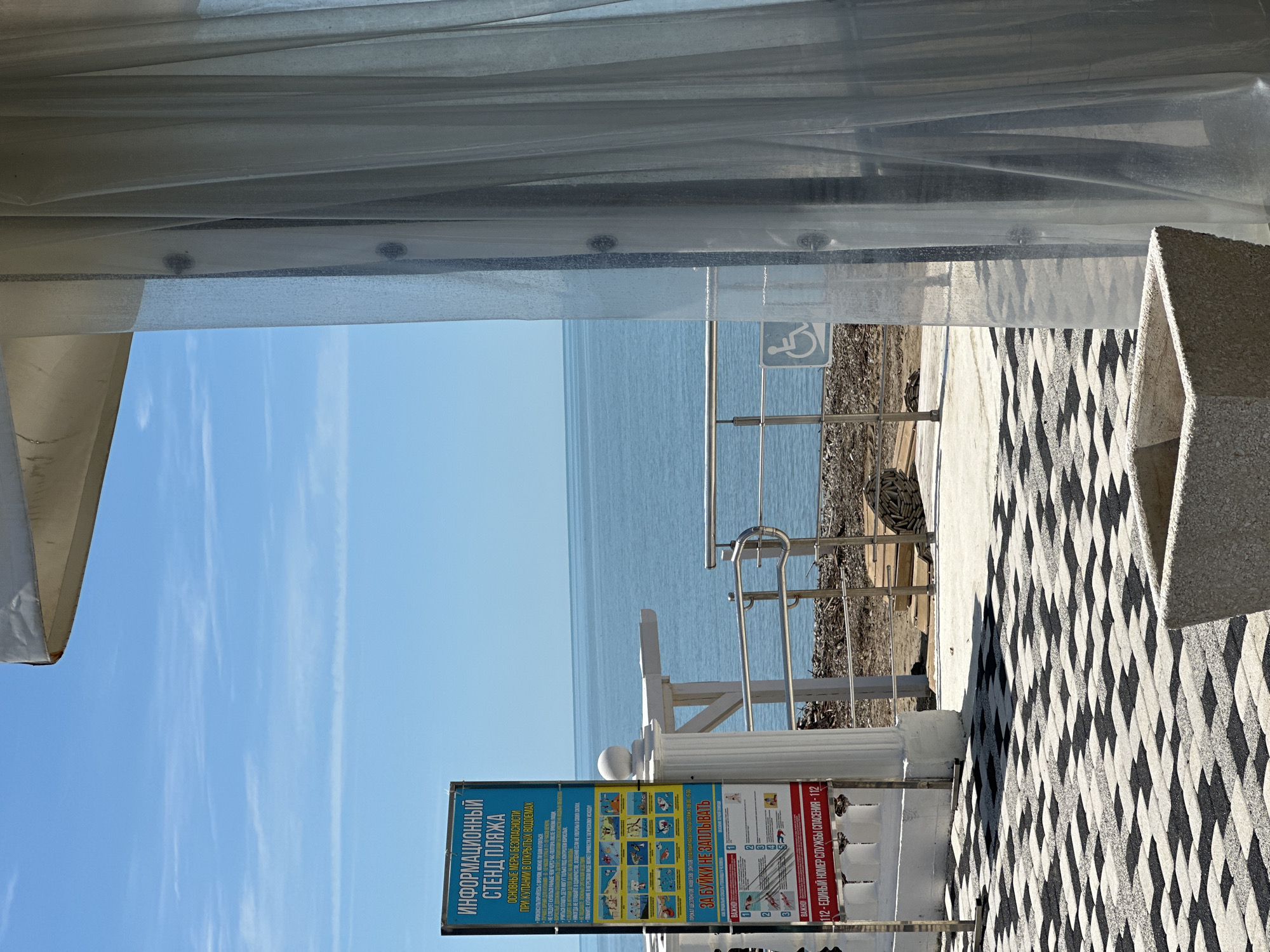 The image, taken in Lermontovo, Russia, captures a bright, sunny scene overlooking a calm sea from what appears to be a sheltered or covered outdoor area. The perspective is from beneath a white canopy or tent, visible along the top and left edge of the frame.
In the foreground, a distinctive black and white geometric patterned pavement covers the ground, leading towards a white decorative pillar and metal railings. To the right, a textured white stone bench or planter is partially visible. A prominent blue informational sign stands upright, titled "ИНФОРМАЦИОННЫЙ СТЕНД ПЛЯЖА" (INFORMATION STAND BEACH). It details "ОСНОВНЫЕ МЕРЫ БЕЗОПАСНОСТИ ПРИ КУПАНИИ В ОТКРЫТЫХ ВОДОЕМАХ" (BASIC SAFETY MEASURES WHEN SWIMMING IN OPEN WATERS). The safety rules include consulting a doctor before swimming, avoiding swimming immediately after eating, supervising children, never swimming alone, and the dangers of swimming while intoxicated. The sign also warns "ЗА БУЙКИ НЕ ЗАПЛЫВАТЬ" (DO NOT SWIM BEYOND THE BUOYS), accompanied by illustrative diagrams. Crucially, it provides emergency contact information: "ВАЖНО! 112 - ЕДИНЫЙ НОМЕР СЛУЖБЫ СПАСЕНИЯ - 112" (IMPORTANT! 112 - UNIFIED EMERGENCY SERVICE NUMBER - 112). Text about renting sun loungers, canopies, and umbrellas, as well as beach operating hours (08:00-19:00), is also present.
Beyond the railings, a sign displaying the international wheelchair accessibility symbol is visible. The ground appears to transition to a natural, dry, and slightly overgrown area with some debris before meeting the water. The serene, light blue expanse of the sea stretches out to the horizon under a clear, bright blue sky dotted with a few faint, wispy clouds. The bright lighting suggests it is daytime, likely morning or early afternoon, with calm and pleasant weather. No people are visible in the scene.