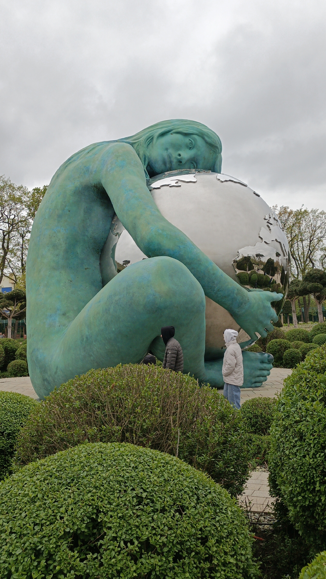 The image captures a view of the "Mother Earth" sculpture in Krasnodar, Russia. The monumental, turquoise-colored sculpture depicts a female figure embracing a globe. Two people stand near the statue; one dressed in a dark jacket with a hood, and the other in a light-colored jacket with a hood, are looking towards the statue. The statue is surrounded by manicured, round bushes in a park setting with trees in the background under an overcast sky.