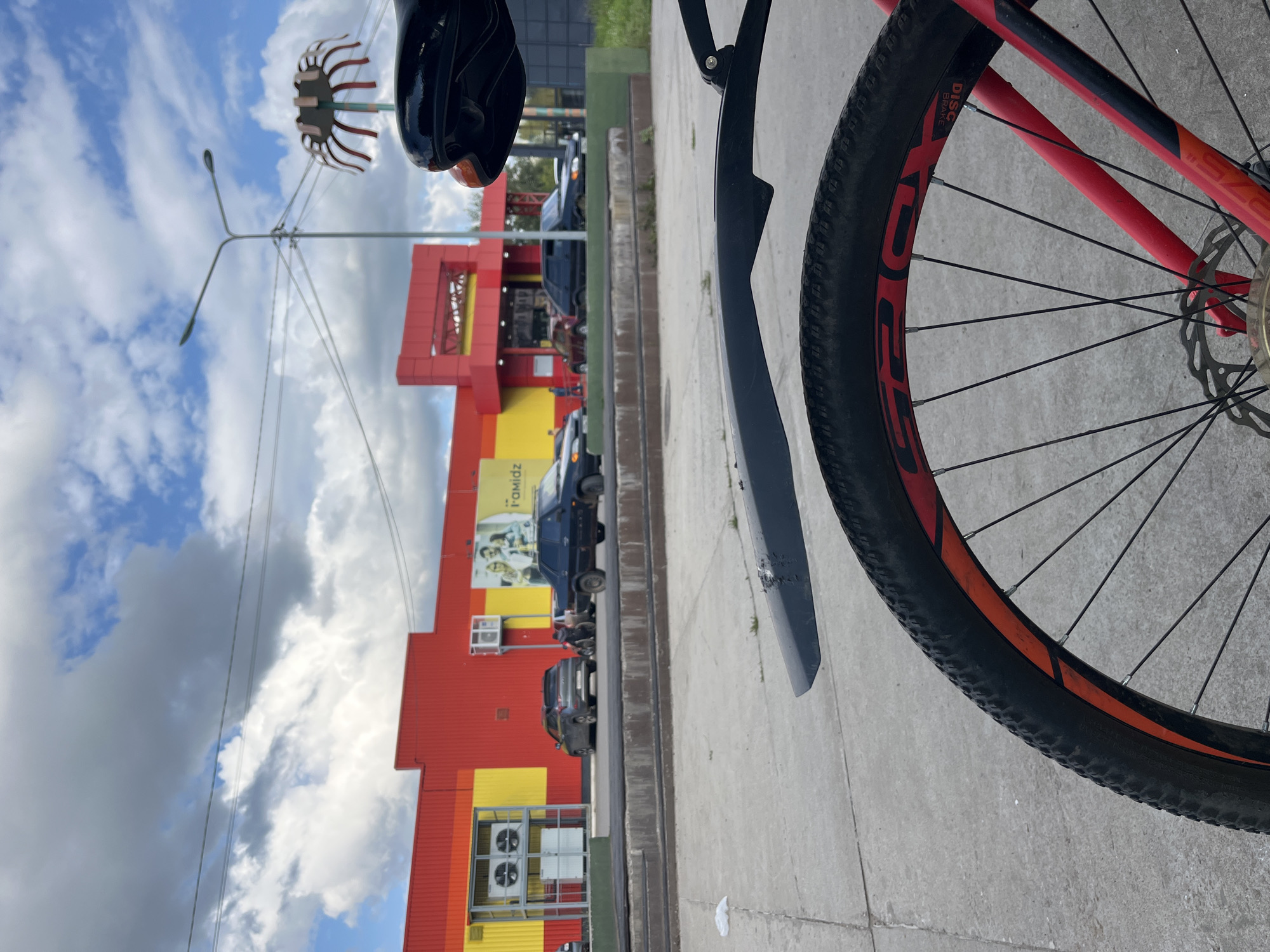 This image, taken in the city of Chaikovsky, Russia, shows a street scene with a red and yellow building in the background that appears to be a retail establishment. In front of the building, there are several cars parked and driving on the road. The foreground of the image is dominated by a close-up view of a bicycle wheel, with part of the frame and fender visible. The sky above is partly cloudy, with a street light and an unusual decorative structure visible. The overall impression is an outdoor urban setting, likely a commercial area, with a focus on a bicycle.