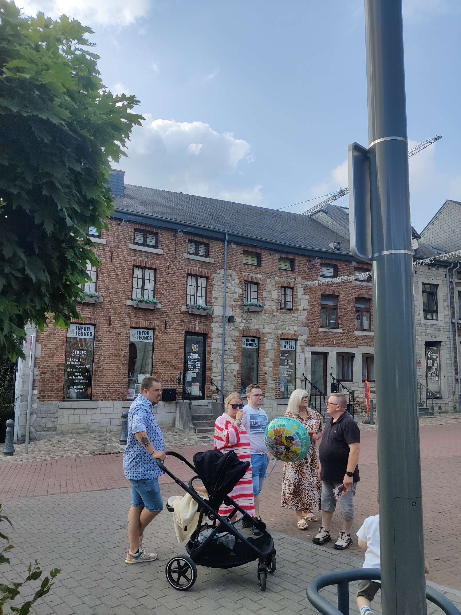 In Marche-en-Famenne, Belgium, on a paved city square, a group of five people stands outdoors. A man in a blue patterned shirt and denim shorts pushes a black stroller. Beside him, a woman in a red and white striped dress and a man in a white t-shirt and denim shorts look towards the stroller. To their right, a woman in a leopard print dress holds a blue balloon featuring cartoon characters, and a man in a dark polo shirt and grey shorts stands next to her. The background features a brick building with signs indicating "Infor Jeunes" and a second building with stone accents. A lamppost is prominent in the foreground, and a crane is visible in the distance, suggesting ongoing construction in the city. The sky is partly cloudy.