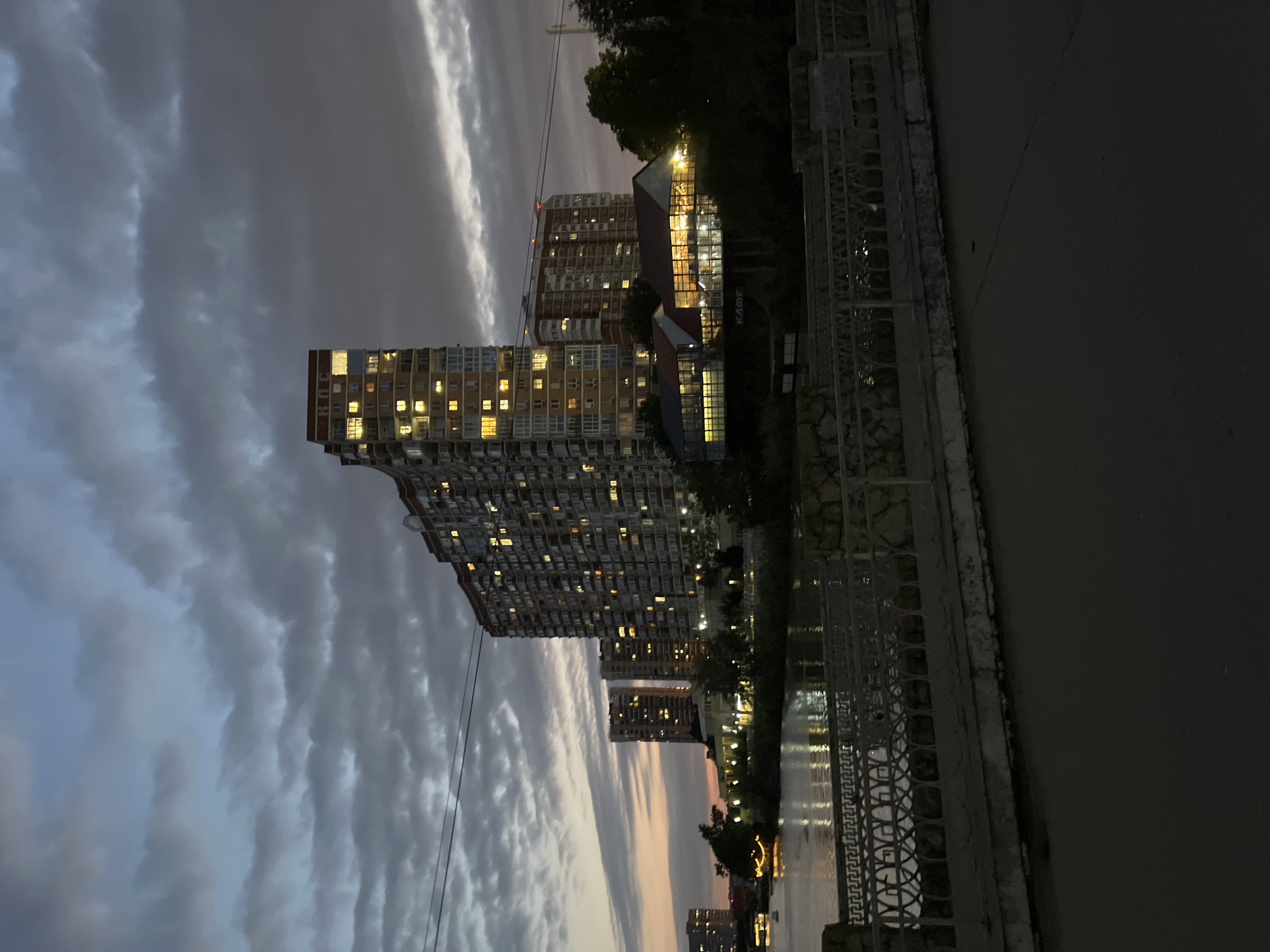 The image depicts a nighttime view of the embankment of the Kuban River in Krasnodar, Russia, near the Bridge of Lovers. Tall, modern apartment buildings with illuminated windows dominate the skyline, reflecting a warm glow onto the rippling water. The sky is filled with dramatic, heavy clouds, transitioning from a soft twilight glow at the horizon to a darker, moody blue overhead.
In the foreground, a pedestrian walkway with a decorative metal railing runs along the riverbank. The railing features an intricate pattern of interlocking shapes and vertical bars. Beyond the railing, the dark, paved surface of what appears to be a street or sidewalk is visible. The river itself is calm, with faint reflections of the city lights and the illuminated buildings creating streaks of light on its surface.
On the far bank of the river, nestled between the apartment buildings and the water, are several low-rise structures. One of these, a modern building with a red roof and large glass windows, is brightly lit, revealing the interior with visible tables and chairs, suggesting it may be a cafe or restaurant. Another building, further to the left, has string lights draped across its facade, adding a festive touch to the scene. Trees and vegetation can be seen interspersed along the embankment, their dark forms silhouetted against the lit buildings and the sky. The overall atmosphere is one of urban twilight, blending natural elements with modern architecture.