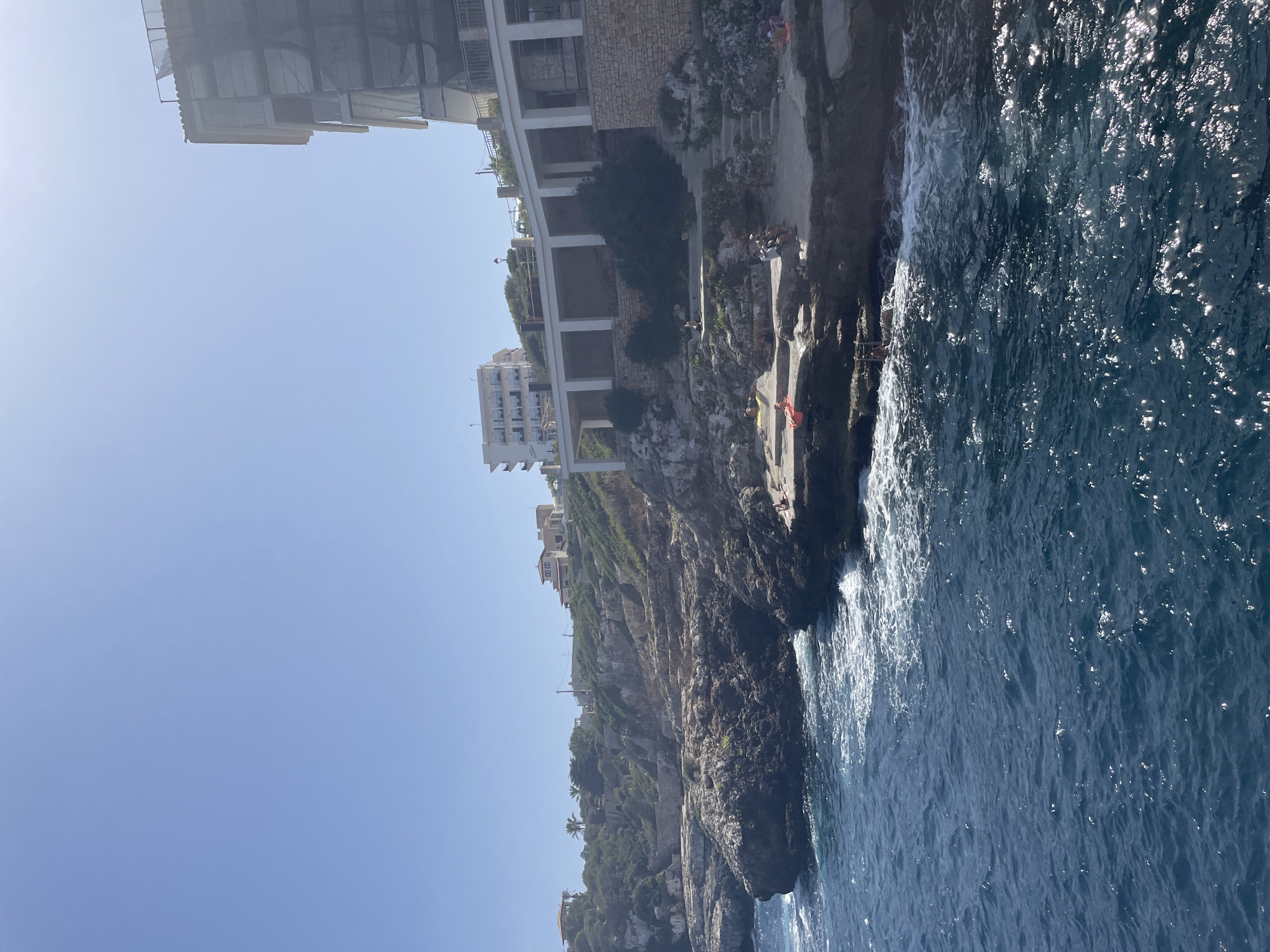 This image depicts a sun-drenched coastal scene, likely in Santanyí, Spain, characterized by rocky cliffs and the deep blue Mediterranean Sea. The foreground is dominated by the choppy, sun-dappled water, with waves crashing against the dark, uneven rocks. Along the coastline, several individuals are visible sunbathing and relaxing on flat rock formations and a small sandy area. One person is lying on an orange float, another pair are seated together, and a few others are scattered, enjoying the seaside.
Above the rocky shoreline, a multi-level structure with white pillars and a white upper deck or promenade is visible. This structure appears to be built into the cliffside. Beyond this, further up the hill, are buildings with light-colored facades, some appearing to be residential or hotel accommodations, interspersed with green foliage and trees. The sky is clear and bright blue. One of the buildings on the right side of the image is partially covered with scaffolding and netting, suggesting ongoing construction or renovation. The overall impression is a popular, sunny spot for leisure and enjoying the coastal environment.
