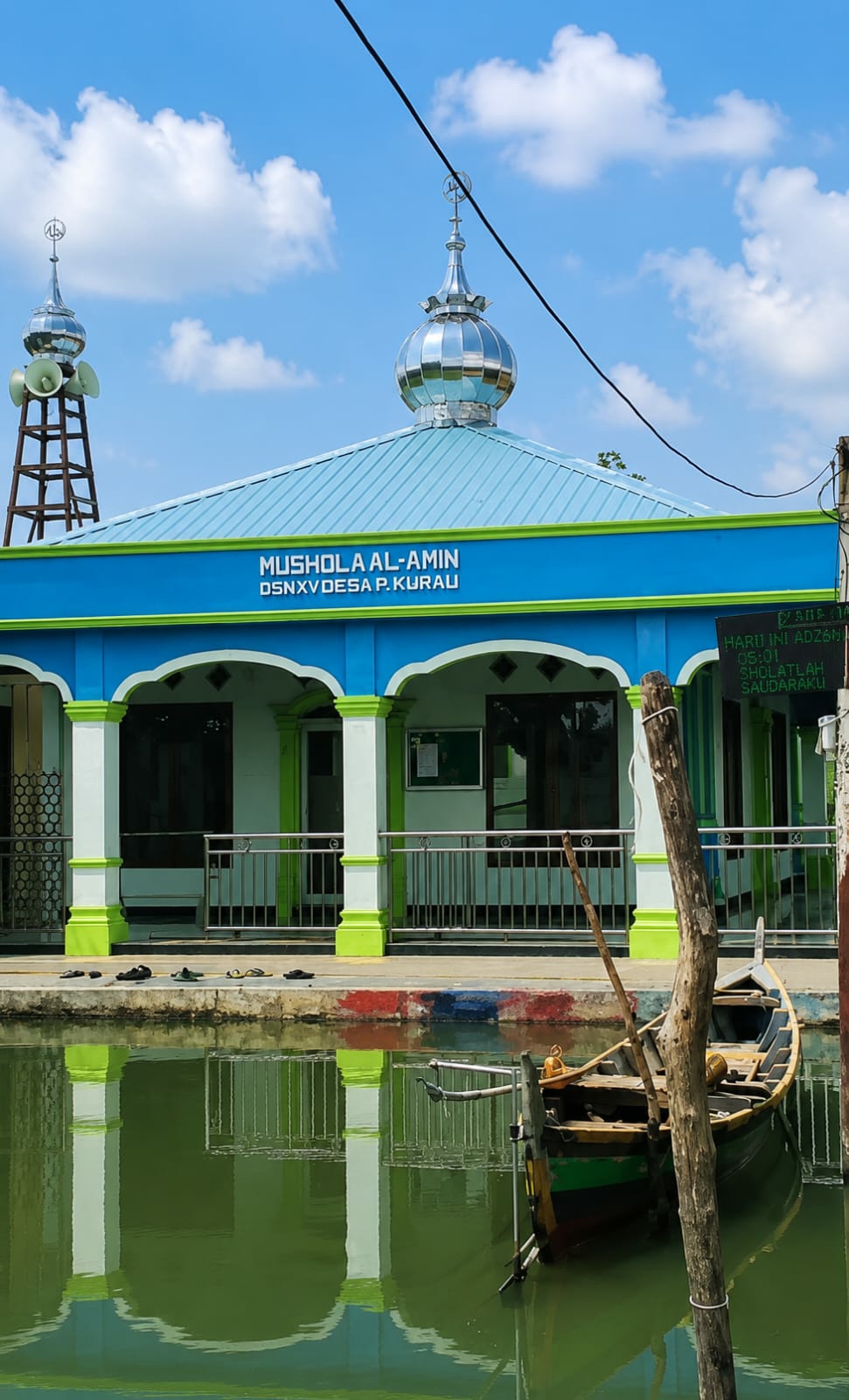 The image shows a mosque with a blue and green facade, a light blue roof, and two domes, one of which has loudspeakers attached to its tower. The mosque is named "MUSHOLA AL-AMIN DSNXV DESA P. KURAU". In front of the mosque, there is a body of water with a small wooden boat. On the right side of the image, there is a digital display showing "HARU INI ADZAN" and a time, followed by "SHOLATLAH SAUDARAKU". The sky is blue with scattered clouds. The image appears to be taken during the day, under good lighting conditions. The presence of a mosque, a boat, and the digital display suggests a potentially coastal or riverine location, possibly in a village area. The mention of "DESA P. KURAU" further supports a rural setting. The overall scene is peaceful and depicts a place of worship and daily life.