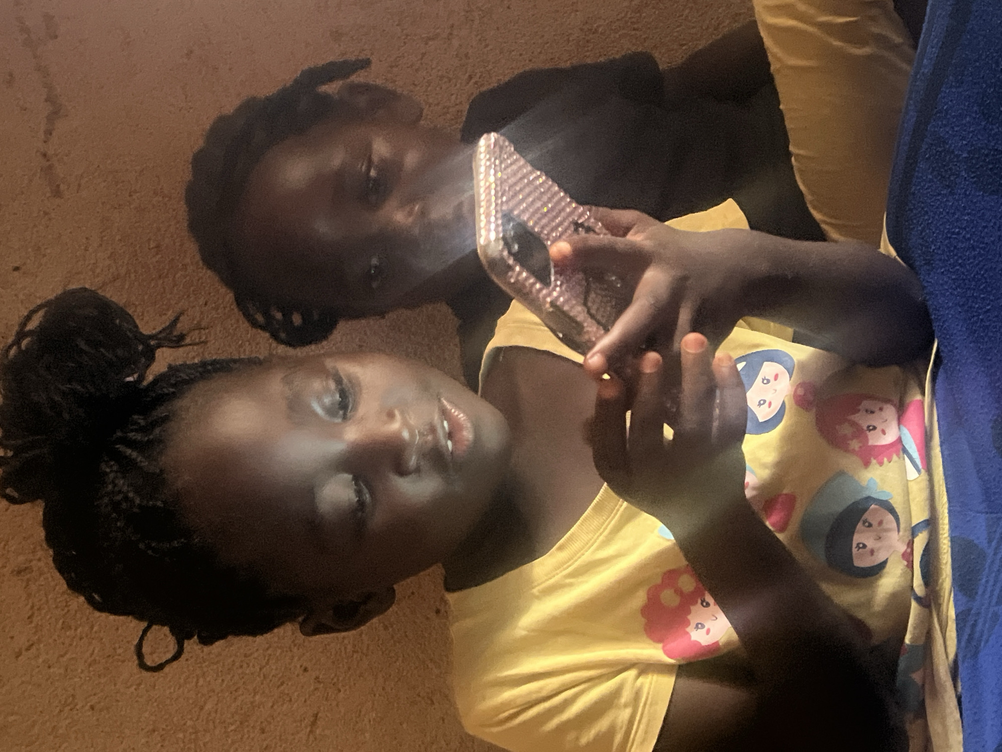 In a dimly lit room with a textured, light brown wall, two young girls are focused on a cell phone. The girl in the foreground, wearing a yellow t-shirt adorned with cartoon characters, holds the phone, which is covered in pink rhinestones. Her gaze is fixed on the screen, and her mouth is slightly open. Behind her, another girl, dressed in a dark shirt, partially obscured, also appears to be looking at the phone. The scene suggests a moment of shared digital engagement in what could be a home environment in Jos, Nigeria.