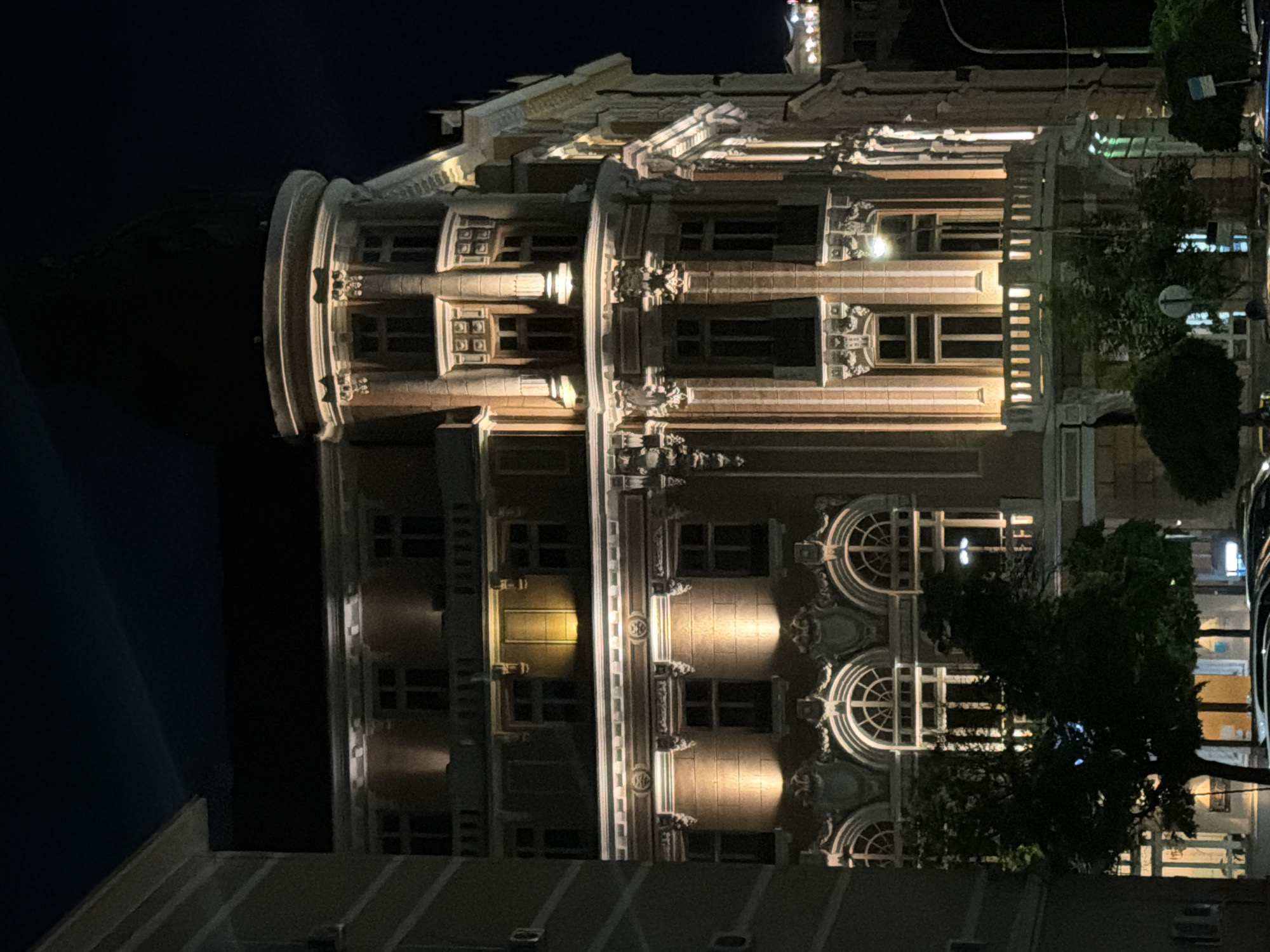 This is a night-time photograph of an ornate building facade illuminated against a dark sky in Sofia, Bulgaria. The building features classical architectural elements, including arched windows on one side, rectangular windows with decorative stone frames on another, and vertical columns separating sections. There are lit windows with light spilling outwards. Trees are partially obscuring one side of the building, and street lights and other buildings are visible in the distance. The photograph is taken from a low angle, looking upwards. The building is the headquarters of the Bulgarian National Bank.