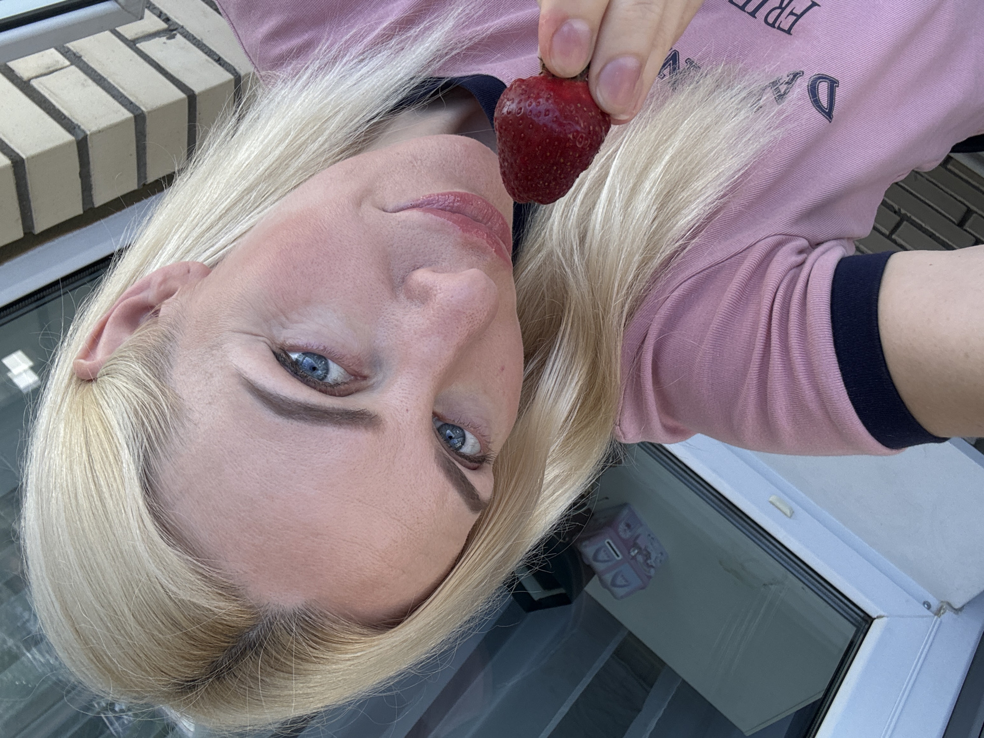 A close-up selfie shows a fair-skinned woman with blonde hair and blue eyes. She is holding a strawberry near her mouth and smiling slightly. The woman is wearing a pink t-shirt with a dark blue trim. The background suggests an outdoor urban setting, possibly a balcony or patio, with a brick wall and a glass door visible. The reflection in the glass shows a pink box with arches. The overall lighting indicates it is daytime.