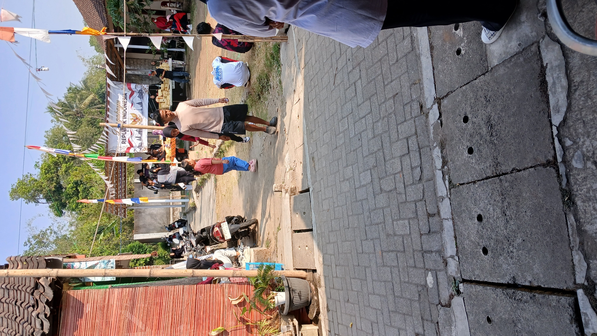 The image depicts an outdoor scene in Mendut, Indonesia, likely during a celebration or event, given the decorations. In the foreground, a paved pathway with concrete drainage covers is visible. Further back, a dirt path leads to a gathering of people. To the left, a red-sided building with a tiled roof is present, next to a potted plant. In the center, a banner reads "REPUBLIK INDONESIA," adorned with Indonesian flags and pennants. Several individuals are seen interacting in the background, including adults and children. Some people are standing, while others are seated. Motorcycles are parked on the side. The environment appears to be a communal outdoor space with vegetation and buildings.