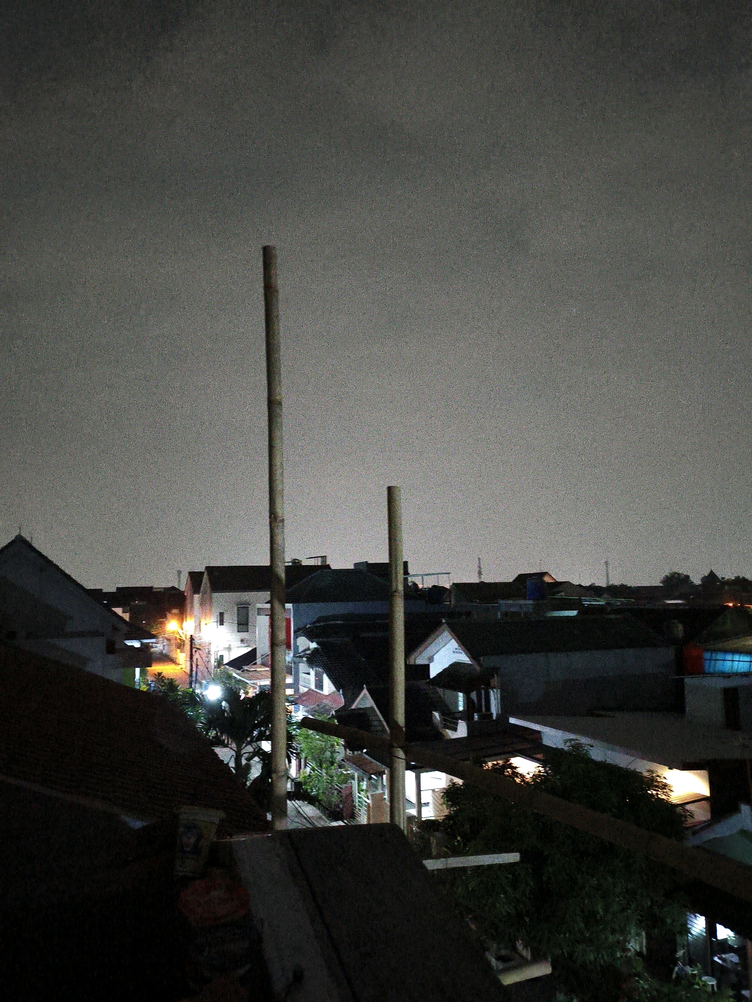 This is a nighttime, medium-distance, eye-level shot of a densely populated urban area, likely in Bekasi, Indonesia, taken from an elevated vantage point, possibly a rooftop. The sky is overcast and dark, with some subtle lighter patches indicating ambient light pollution.
Dominating the foreground are the dark, angled roofs of houses. Two tall, thin wooden poles stand prominently in the mid-ground, positioned vertically and slightly off-center. One pole is in the left half of the frame, and the other is in the right half. Between and behind the poles, a cluster of houses and buildings are visible.
These structures exhibit a variety of architectural styles, with some featuring illuminated windows that cast a warm, yellowish glow onto the surroundings. One building in the center appears to be a multi-story dwelling with white walls and dark rooflines, brightly lit from within. Other buildings have dark facades, making them blend into the night. There are streetlights visible, casting pools of light on what appear to be pathways or small roads winding through the settlement.
Trees and vegetation are interspersed among the buildings, adding a touch of green to the otherwise monochromatic scene. The overall impression is of a bustling, lived-in urban environment at night, characterized by the interplay of artificial light and darkness. There are no people or specific actions depicted in the image. The image quality is somewhat grainy, consistent with a low-light photograph.