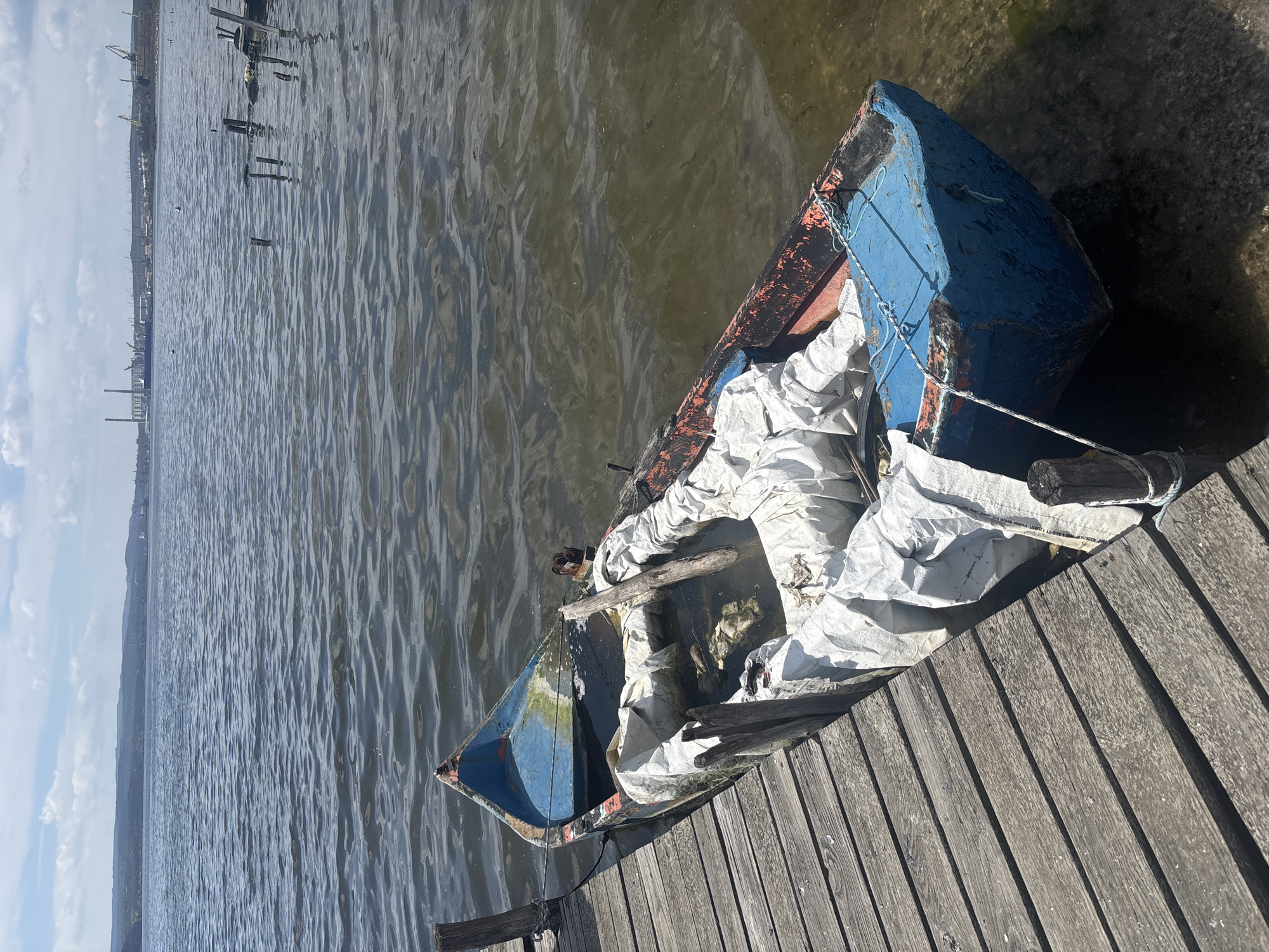The image depicts a dilapidated, blue and orange rowboat partially sunk and lying on a wooden dock, partially submerged in water. The boat is filled with white, possibly plastic, bags and miscellaneous debris. The dock is constructed of weathered wooden planks. In the background, water stretches towards a distant shoreline marked by indistinct industrial structures and a hazy sky. The image was taken in or near Казашко, Bulgaria.