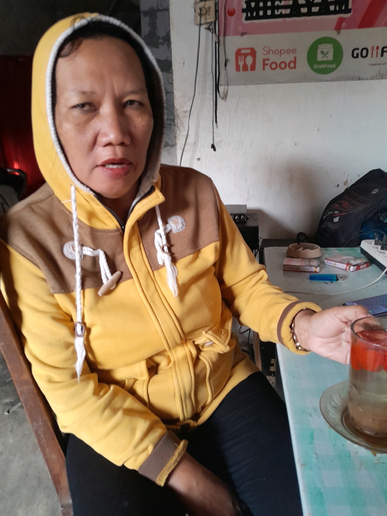 A woman is seated at a table in what appears to be a humble establishment in Glanggang, Indonesia. She is wearing a yellow hooded jacket with brown accents and black pants. In front of her, on the table, are a glass of liquid, a small ashtray, cigarette packs, a lighter, and a mobile phone. The background reveals a wall with some posters, including one with the Shopee Food and GrabFood logos, suggesting a connection to food delivery services. The overall scene depicts a casual moment, possibly a break or a wait, in a local setting.