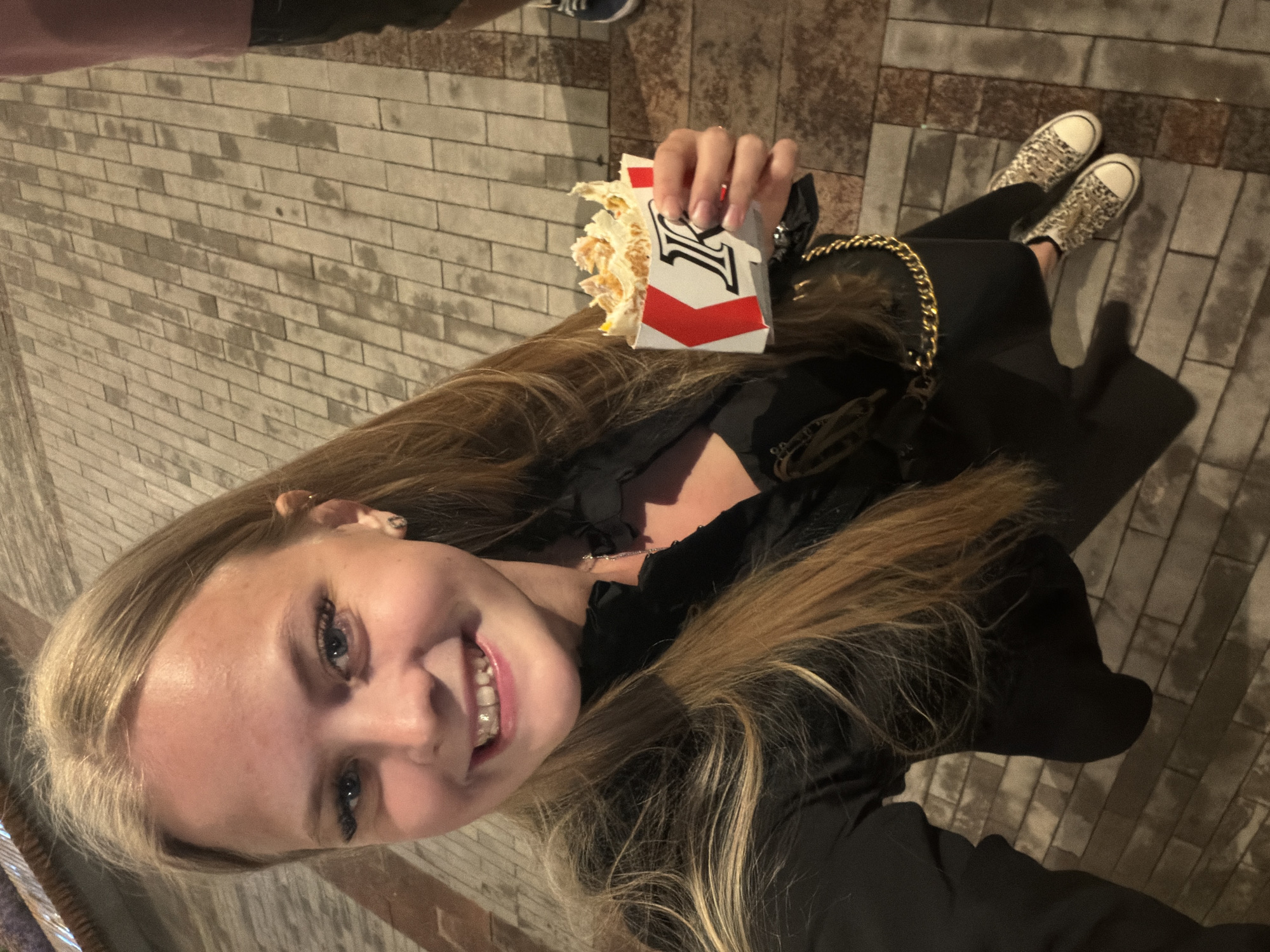 A young woman with long blonde hair smiles at the camera, holding a partially eaten wrap in her right hand. She is wearing a black jacket over a black top, a gold chain detail on her bag, and leopard print sneakers. The background is a cobblestone sidewalk, typical of a city street. There is no specific indication in the image to suggest the location is Pervomaysky, Russia.
