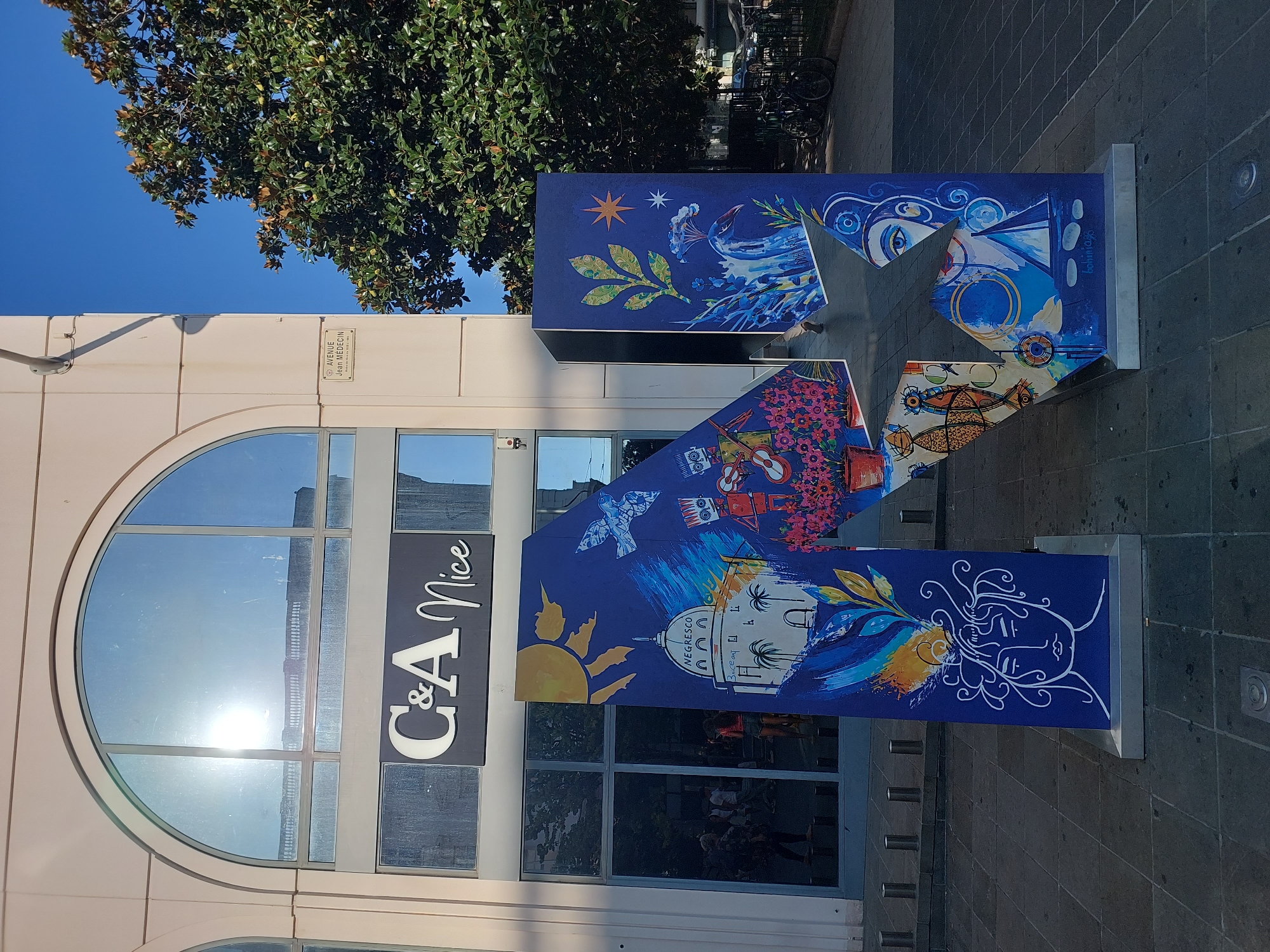 The image shows a large, blue, three-dimensional letter "N" sculpture adorned with various paintings and designs, standing on a paved sidewalk in front of a building with arched windows and a "C&A Nice" sign. A street sign that reads "AVENUE Jean Médecin" is visible on the building's facade. To the right of the sculpture is a large tree with green leaves. The sculpture itself features diverse artistic elements, including a depiction of the Negresco hotel, palm trees, a sun, a bird, flowers, a face, and abstract patterns. The sky is clear and blue. The scene appears to be in a commercial area of Nice, France, specifically on Avenue Jean Médecin, a well-known shopping street.