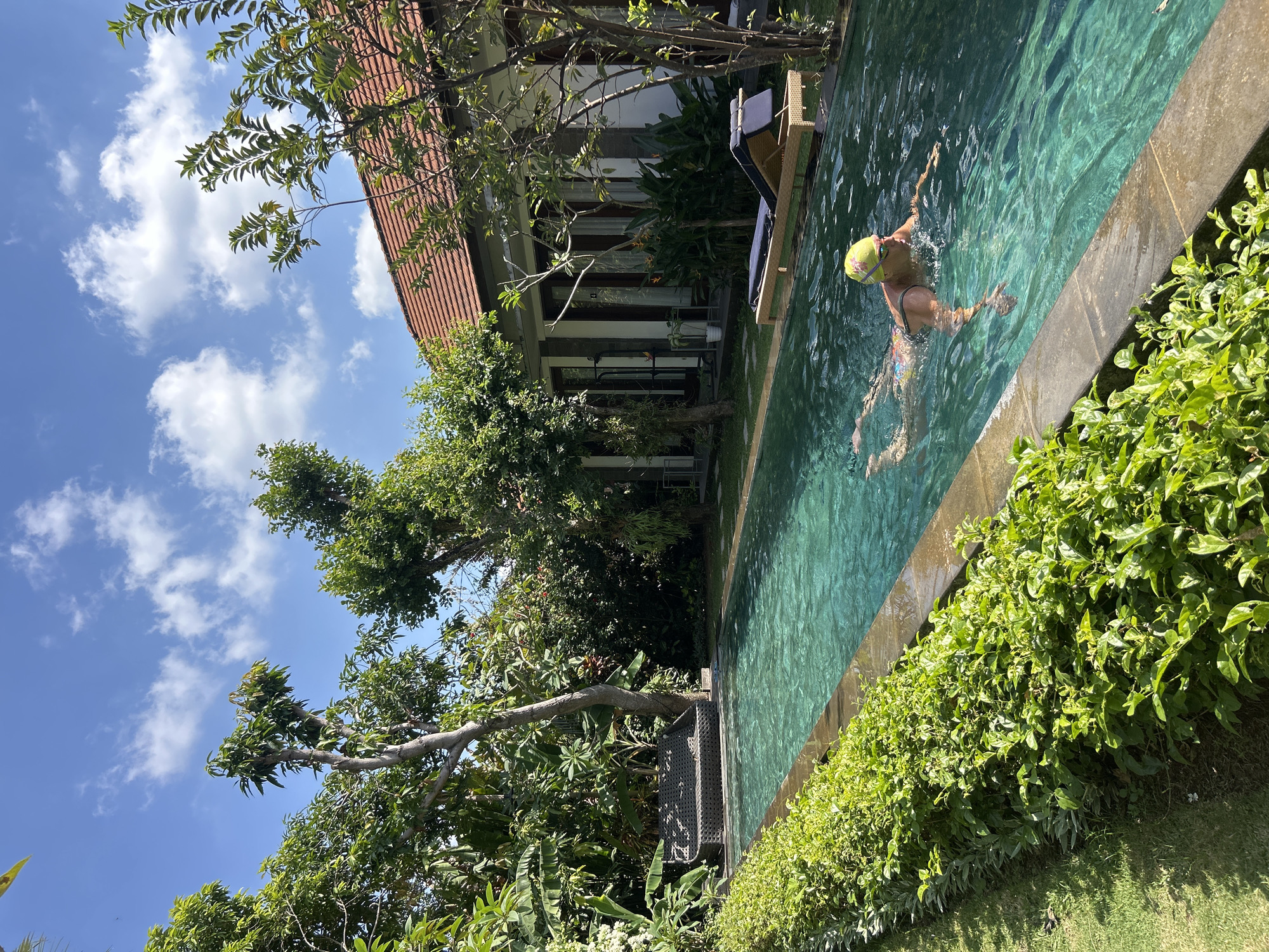 In the city of North Kuta, Indonesia, a person wearing a yellow swim cap and goggles swims in a rectangular swimming pool. The pool is lined with lush green bushes and has a stone edge. To the right of the pool, a building with a red tiled roof and white walls is visible, with several doorways and windows. A lounge chair with a blue cushion sits on a wooden platform near the building. The area is surrounded by abundant green foliage and tall trees, and the sky is bright blue with scattered white clouds.