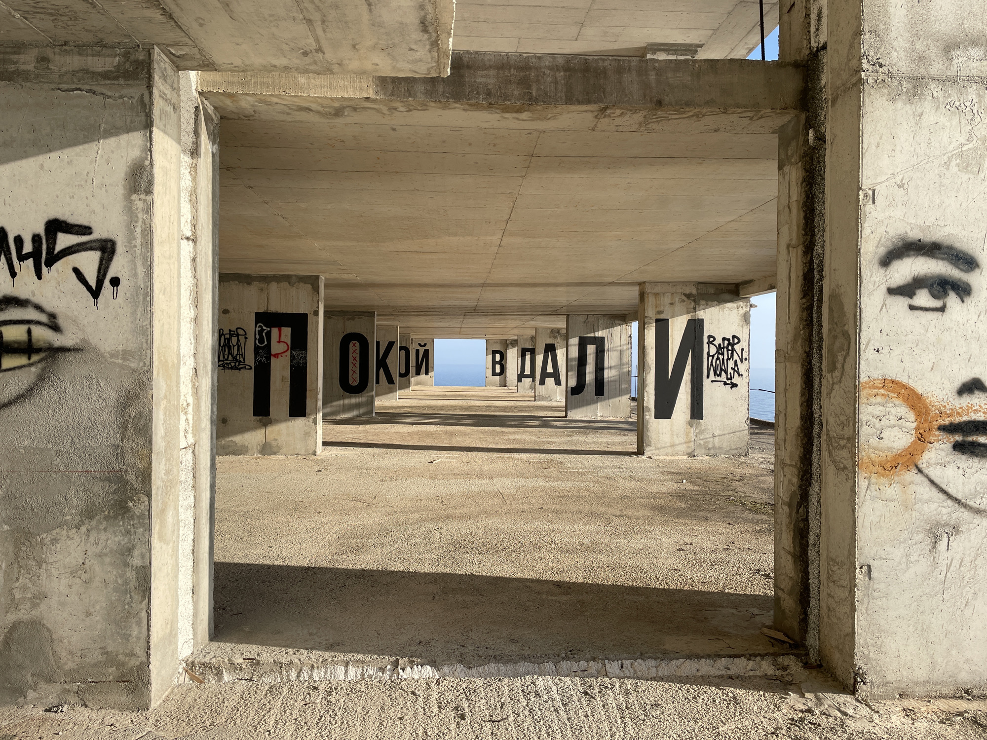 The image captures an interior perspective within a concrete structure located in Perazića Do, Montenegro. The space is defined by a series of concrete pillars and ceilings, forming a linear corridor. Each pillar is marked with black graffiti, spelling out words in Cyrillic script. Additional graffiti decorates the pillars framing the foreground. The floor is covered in a layer of light-colored sediment. Light streams through the open end of the structure, illuminating a glimpse of the sea and sky in the distance. The structure appears unfinished or abandoned.