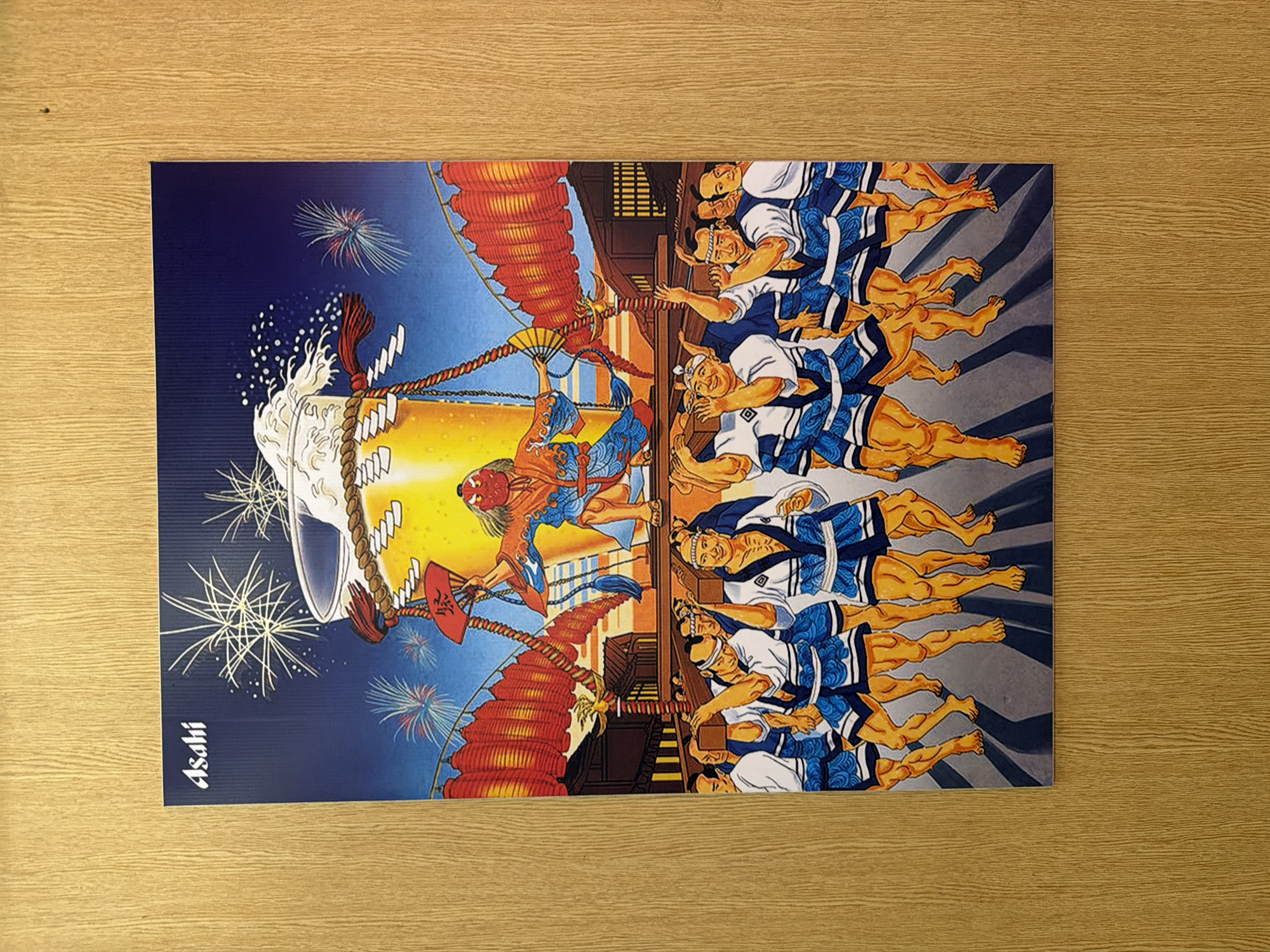 The image is a vibrant, stylized illustration depicting a Japanese festival scene. On the right side, a row of eight muscular men is shown in dynamic poses, dressed in traditional white shirts, blue and white patterned shorts or loincloths, and matching headbands. Their bare, tanned legs and arms are prominently featured, and their expressions are joyful and energetic.
To the left of the men, a large, ornate float is depicted, appearing to be pulled or celebrated. On the float, a central figure with a red demon-like mask and colorful blue and orange robes holds a fan. The float itself features multiple tiers of red, stacked elements, possibly lanterns or decorative roofs, and is adorned with thick ropes and white paper streamers (*shide*). A large, stylized white foamy element, possibly representing water or a cloud, is positioned at the top center of the illustration.
The background is a dark blue night sky, illuminated by scattered white and colorful fireworks. A strong yellow-orange light glows from the central background, suggesting the illumination from lanterns or a powerful central light source. Traditional Japanese-style buildings are partially visible in the background behind the men.
The activity taking place is a lively festival procession, with the men enthusiastically participating in pulling or celebrating around the elaborate float. The overall atmosphere is one of communal celebration, excitement, and energy. The artwork employs bold colors and exaggerated details, particularly the musculature. The word "Asahi" is written vertically in white, stylized script on the far left side of the image.