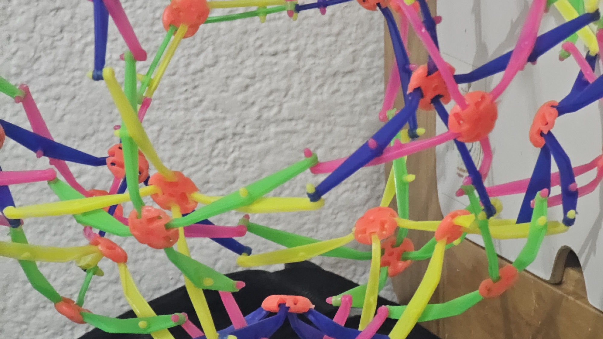 A close-up shot shows a complex spherical structure made of colorful plastic rods and orange connectors. The rods are predominantly blue, green, yellow, and pink. The structure is resting on a dark surface. The background is out of focus, revealing a textured white wall and a portion of a wooden surface. San Bernardino Tlaxcalancingo, Mexico is not evident from the provided image.