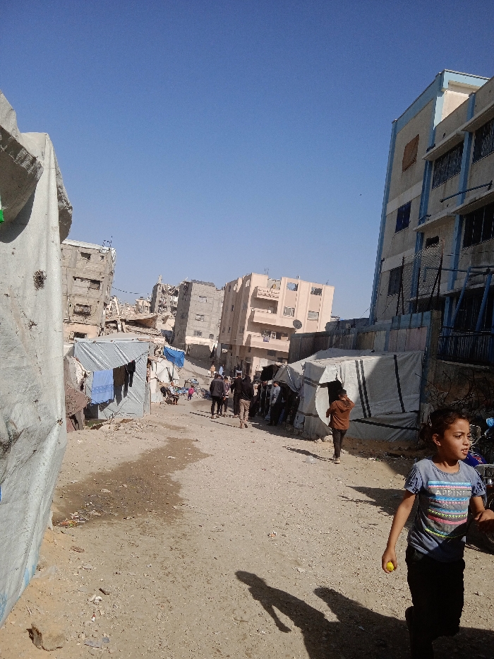 The image displays a daytime scene along an unpaved, dusty street in Khan Yunis, Palestinian Territory. In the foreground, a young girl with dark hair, wearing a blue graphic t-shirt and dark pants, walks towards the right, holding a small yellow object in her right hand. Her elongated shadow is cast on the ground. To the left of the girl, several individuals, mostly adults dressed in dark clothing, walk away from the viewer along the center of the path.
On the left side of the frame, a large, light-colored fabric tent structure occupies the immediate foreground. Beyond it, multiple makeshift shelters constructed from corrugated metal sheets and tarps line the street, with blue laundry hanging from a line in front of one. The dirt ground exhibits damp patches and scattered debris.
In the mid-ground and background, numerous multi-story buildings show signs of damage, including visible structural deterioration, exposed concrete, and missing sections. A beige-colored multi-story building is prominent in the center-right background, and a taller building with blue trim and multiple windows is visible on the far right. The sky overhead is clear and blue.